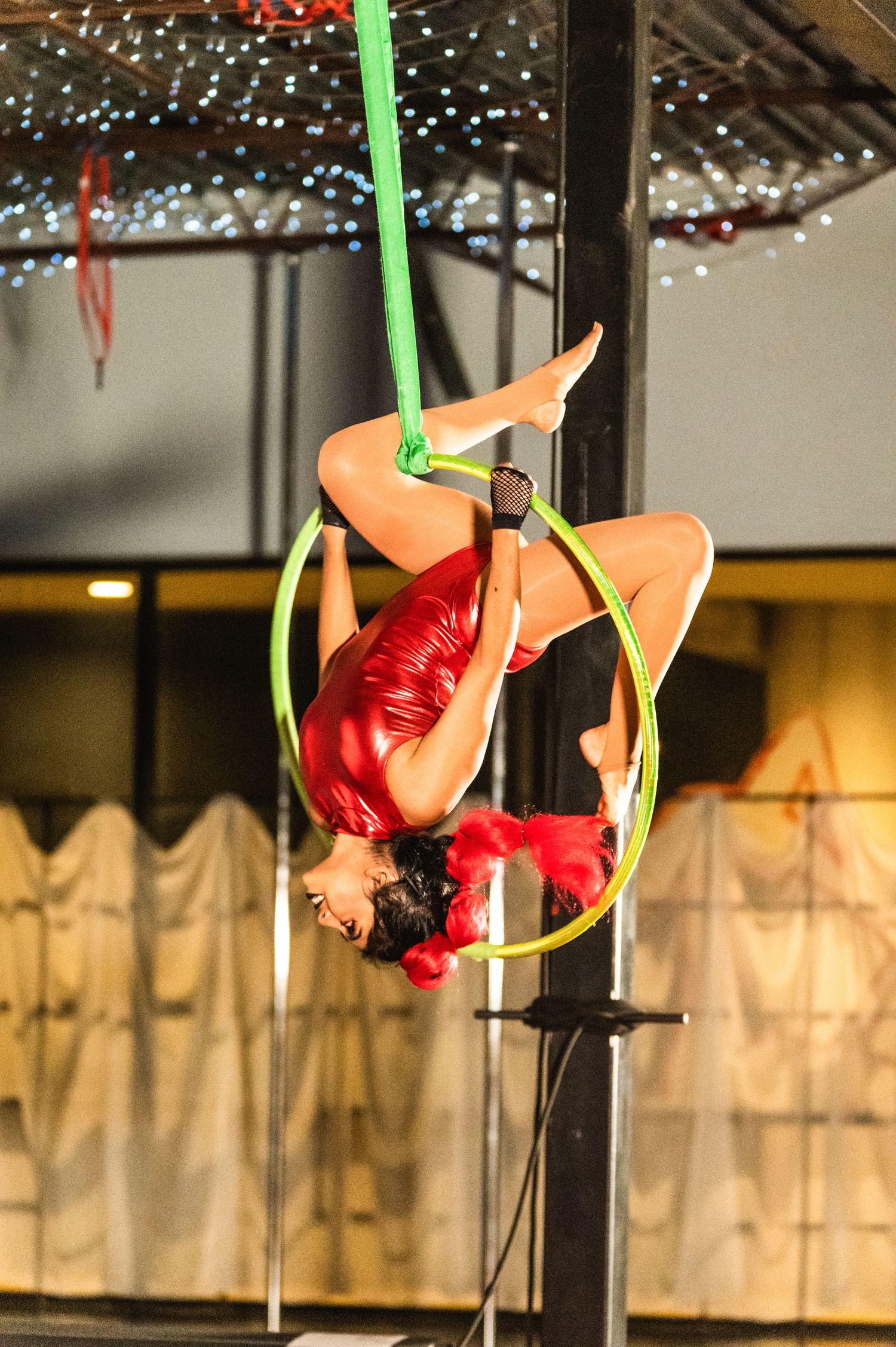 A performer in red attire executes an aerial maneuver on a green hoop under twinkling lights.