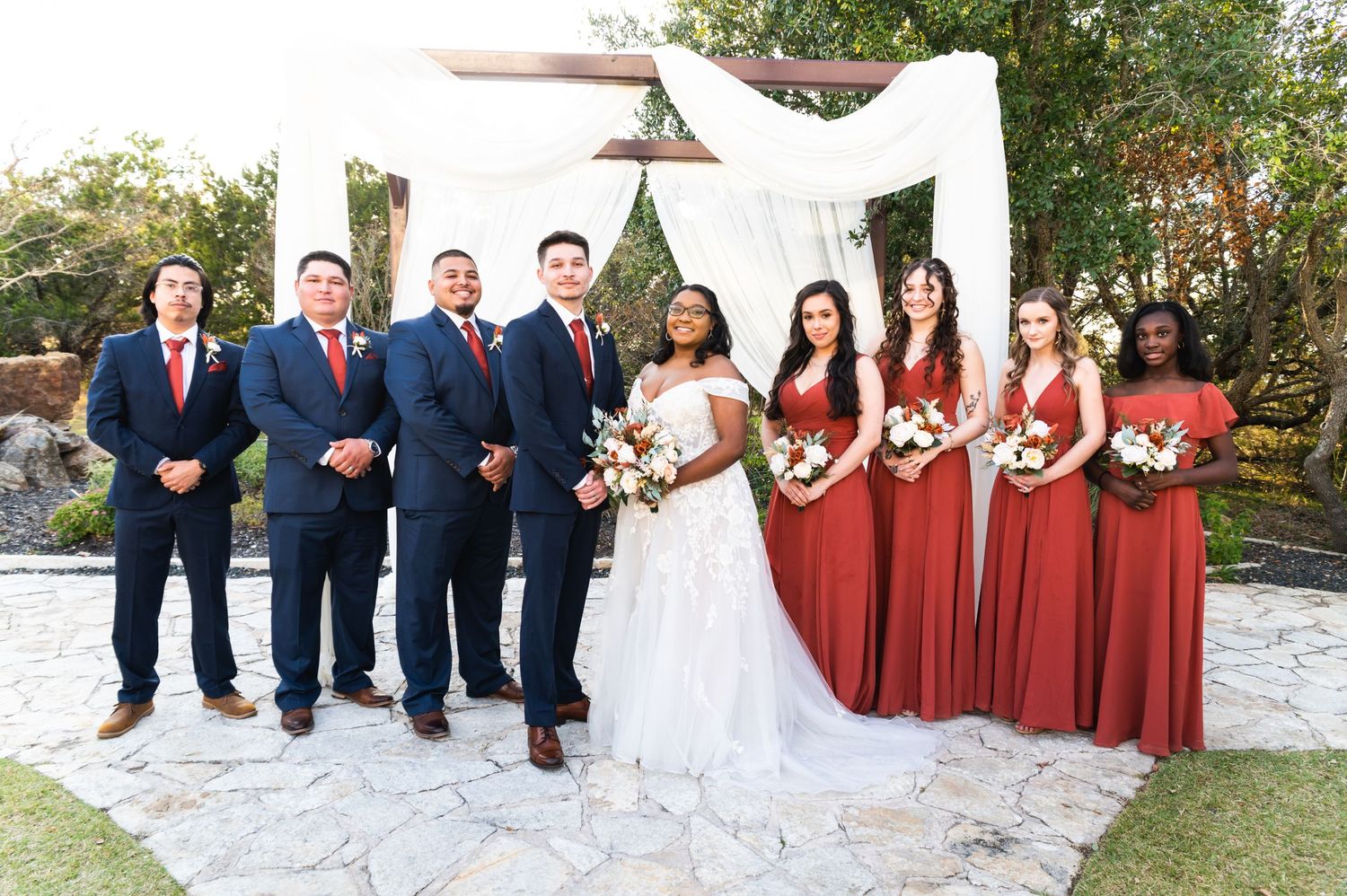 Wedding party in navy suits and rust-colored dresses pose under a white draped arch outdoors.