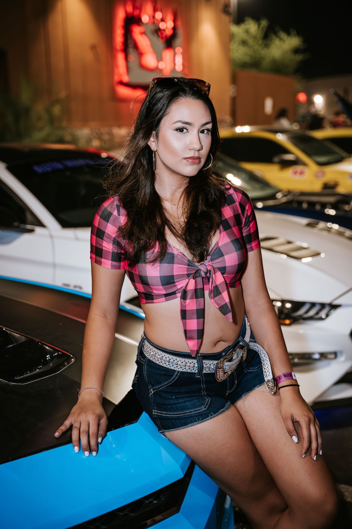 Model in pink plaid top sits on blue sports car hood at night car meet.