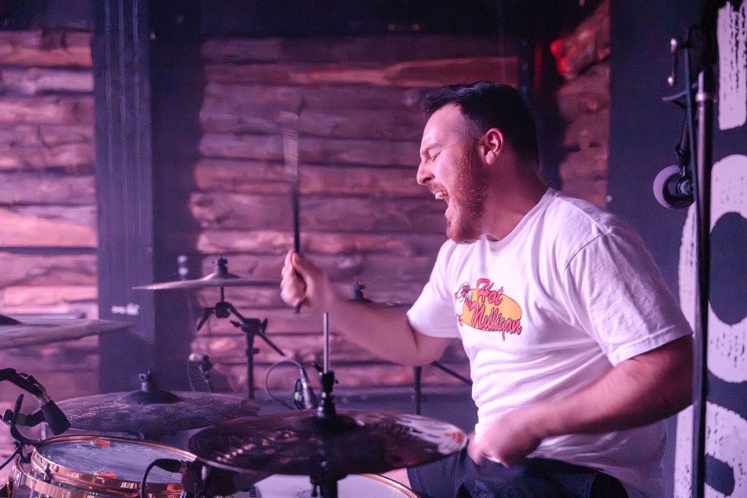A drummer plays energetically on a drum kit in a rustic venue with wood-paneled walls.