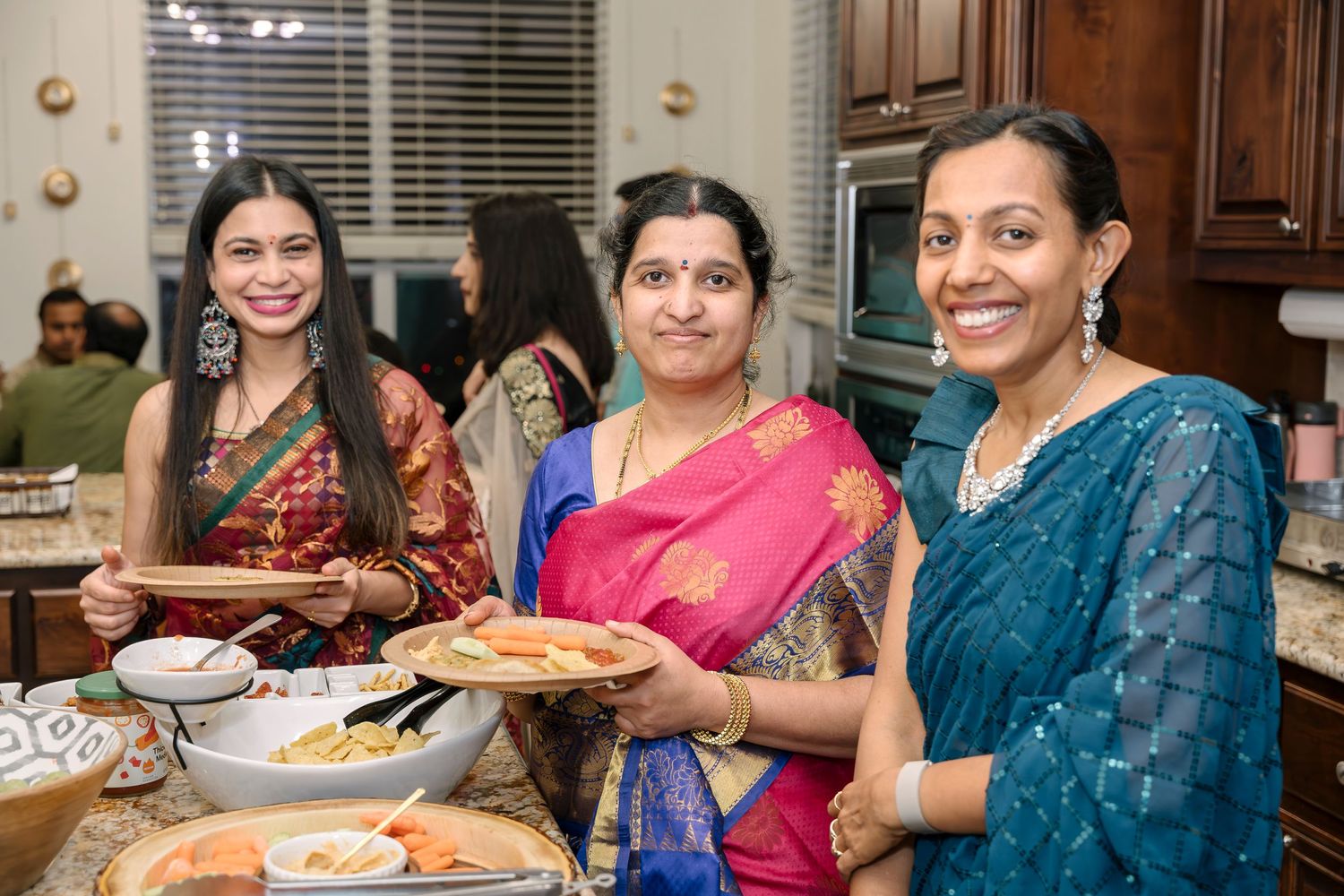 Three women in colorful sarees serving food at an indoor gathering.