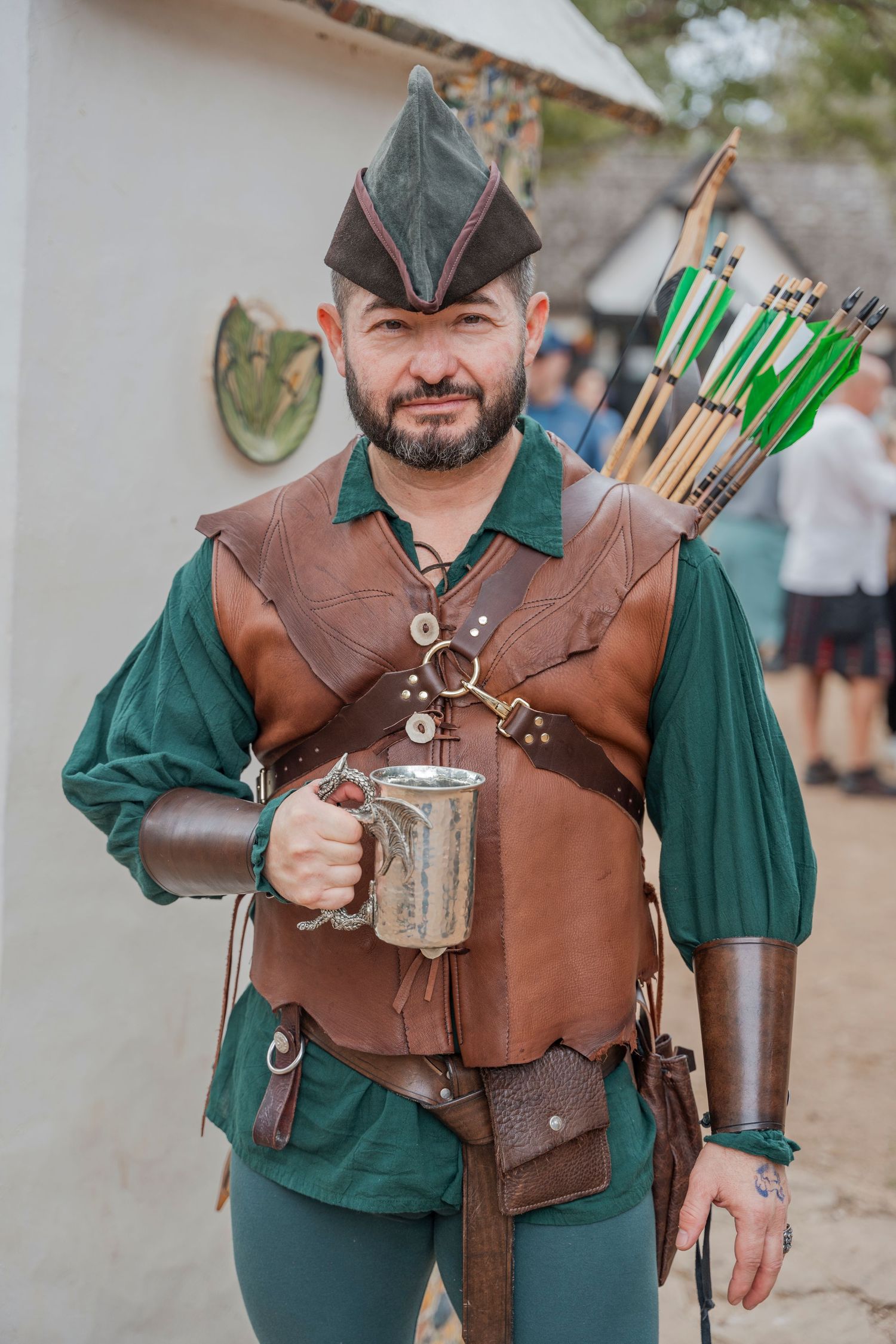 Archer in medieval costume with leather vest and green tunic holding a tankard.