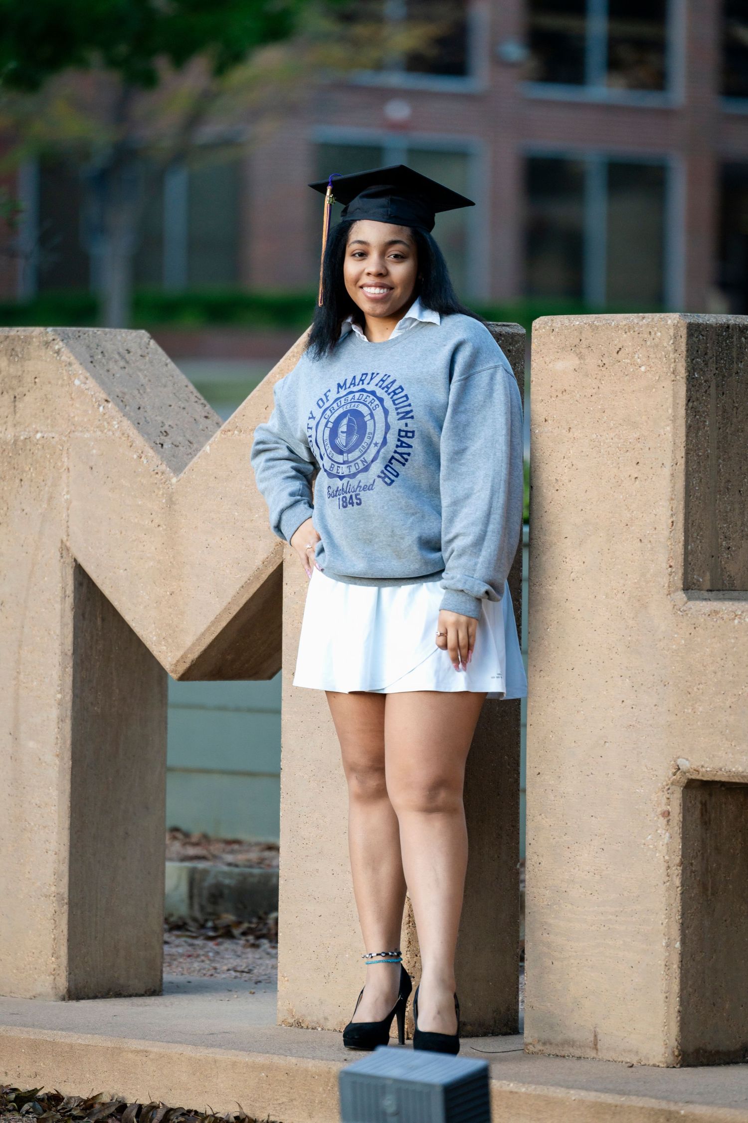 Graduate wearing cap and grey university sweatshirt poses on concrete UMHB sign.