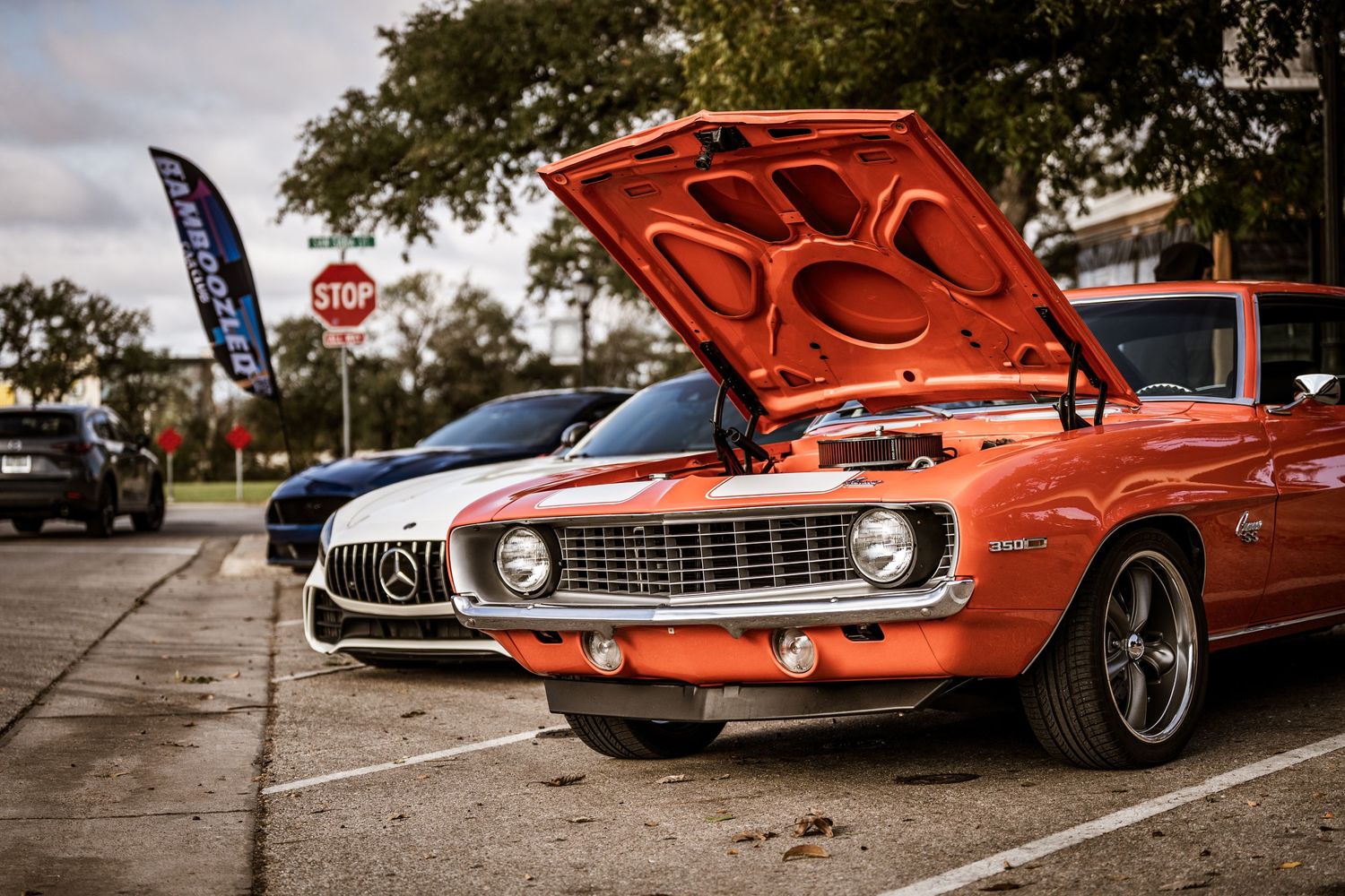 Classic orange Camaro with hood open displayed at a car show next to modern Mercedes.