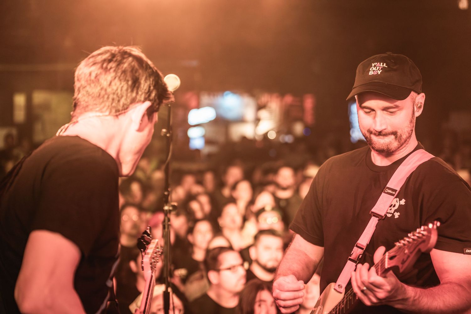 Two guitarists perform on stage at a crowded music venue with warm lighting and audience in background.