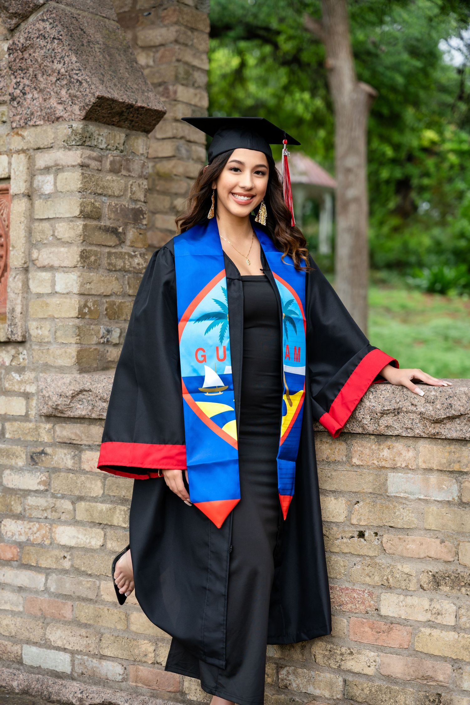 Graduate in cap and gown with decorative stoles stands against brick wall surrounded by greenery.