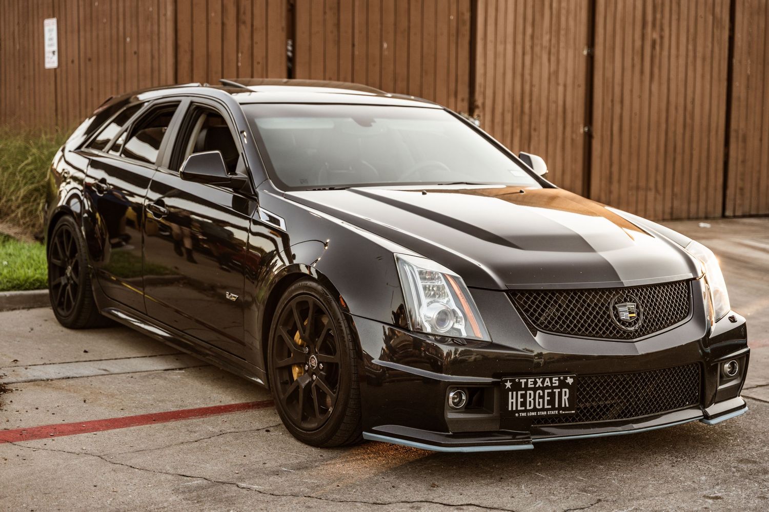 Black Cadillac CTS-V with custom wheels and modifications parked against a wooden fence.
