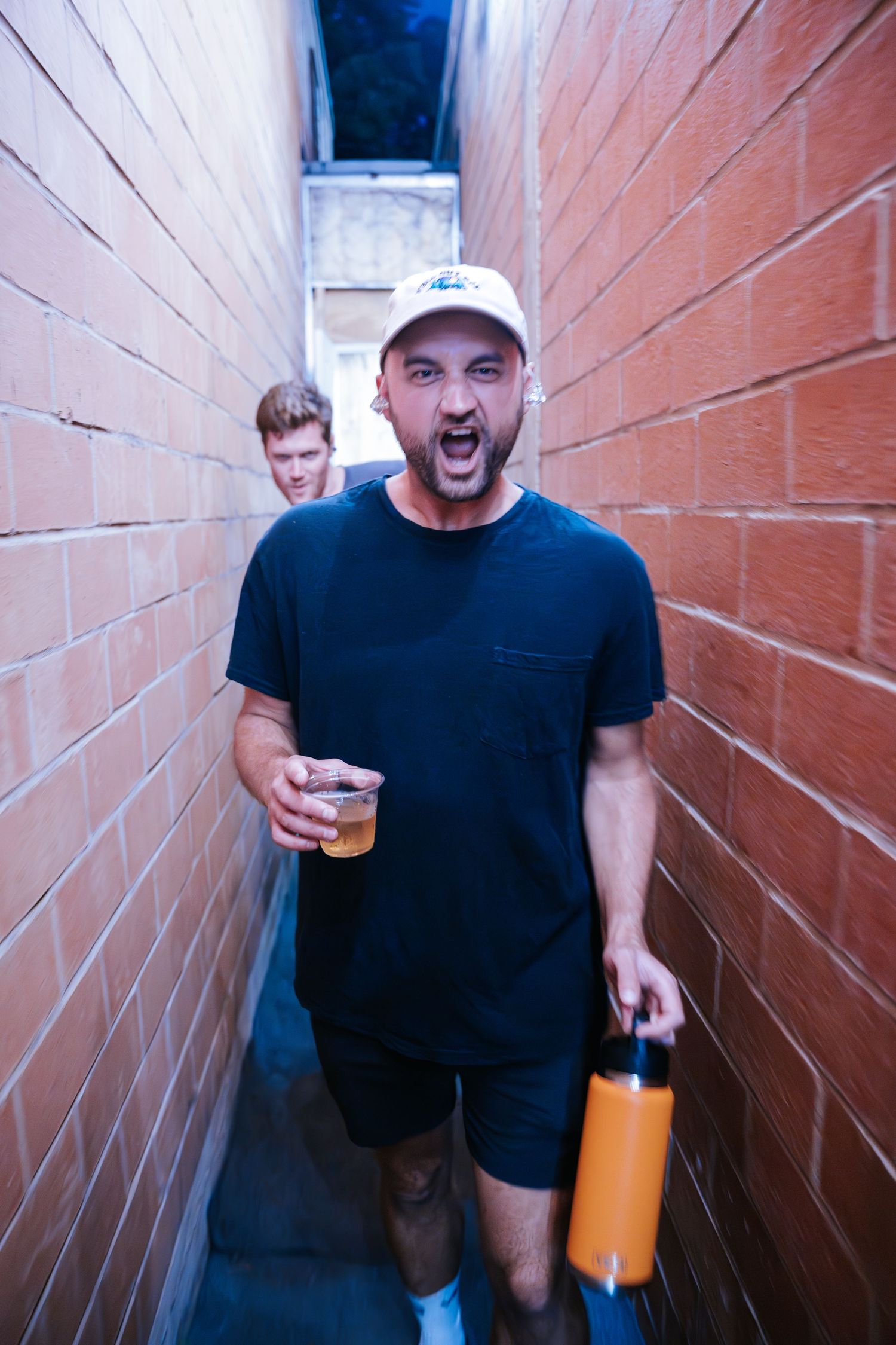 Person energetically holding a drink and orange bottle in a narrow brick alleyway.
