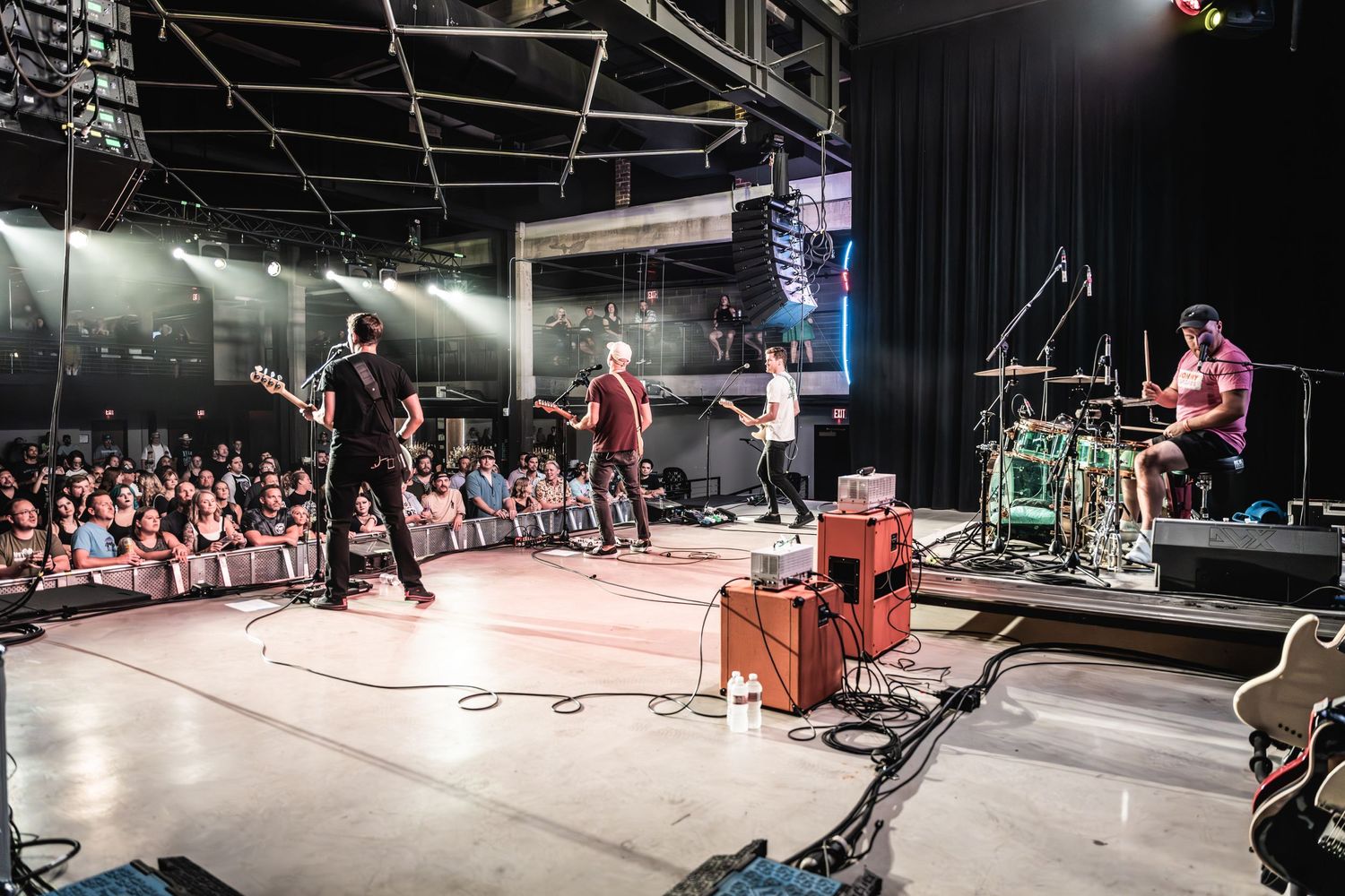 Wide shot of band performing on stage in front of large crowd at indoor venue.