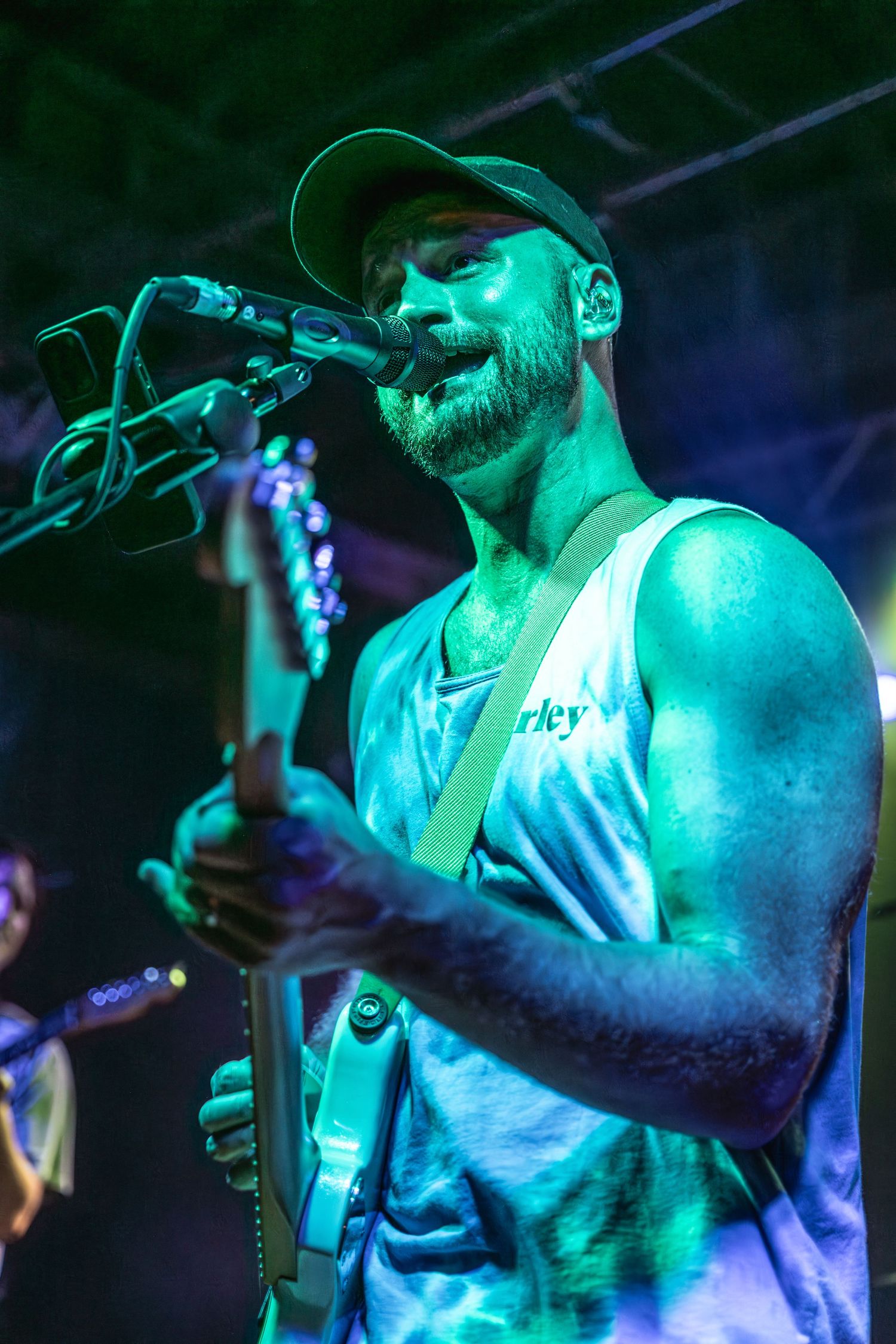Musician performing on stage bathed in teal lighting while singing into a microphone wearing a baseball cap.
