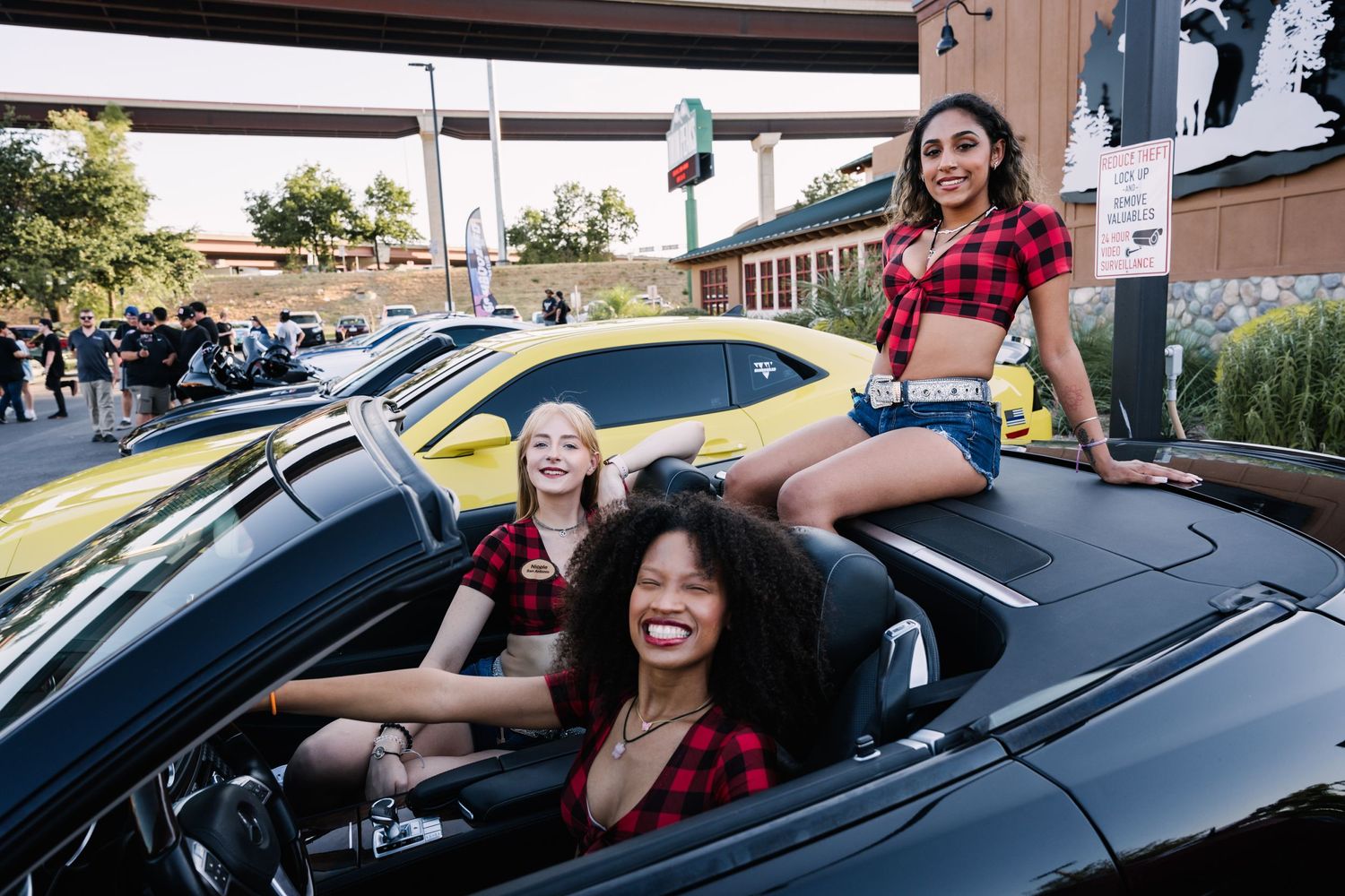 Models in matching red plaid shirts pose together in and on a black convertible sports car.