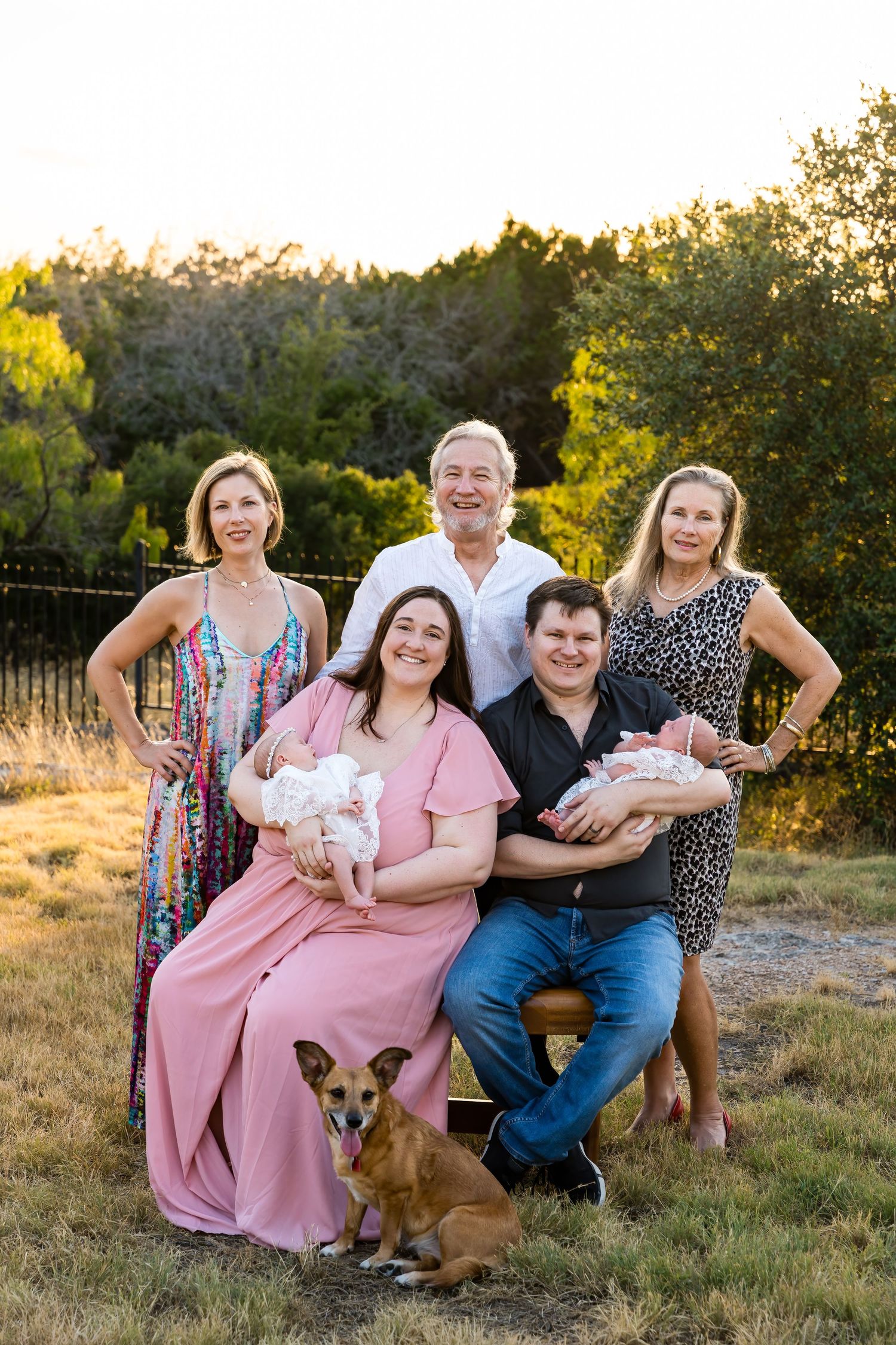 Multi-generational family portrait outdoors with twin babies and a corgi dog at sunset.