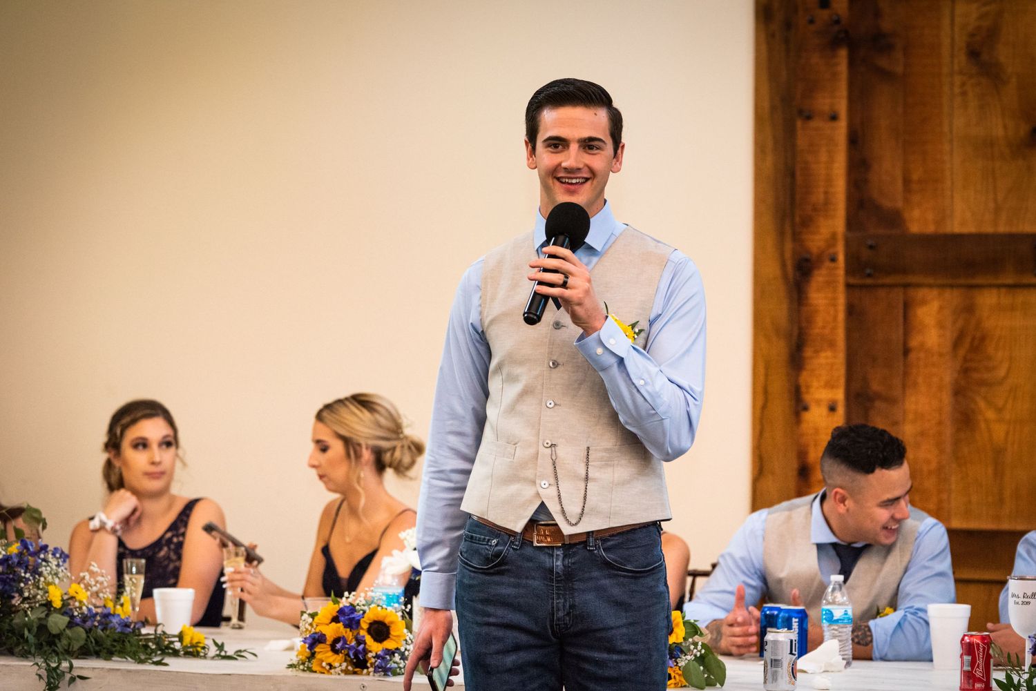 Person giving a wedding speech at reception with rustic barn doors and floral table decorations.