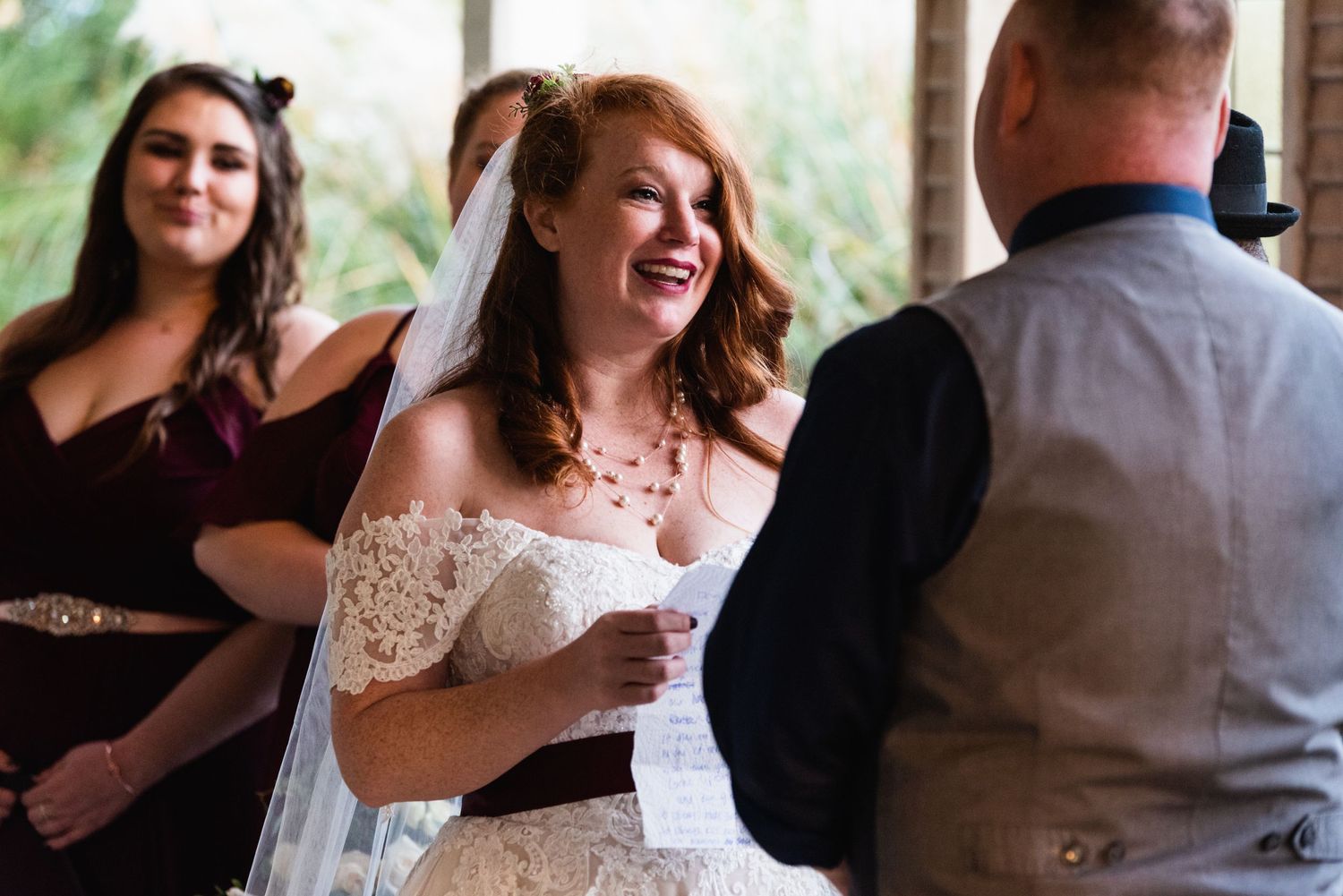 Candid moment of laughter during wedding ceremony with burgundy-clad bridesmaid in background.