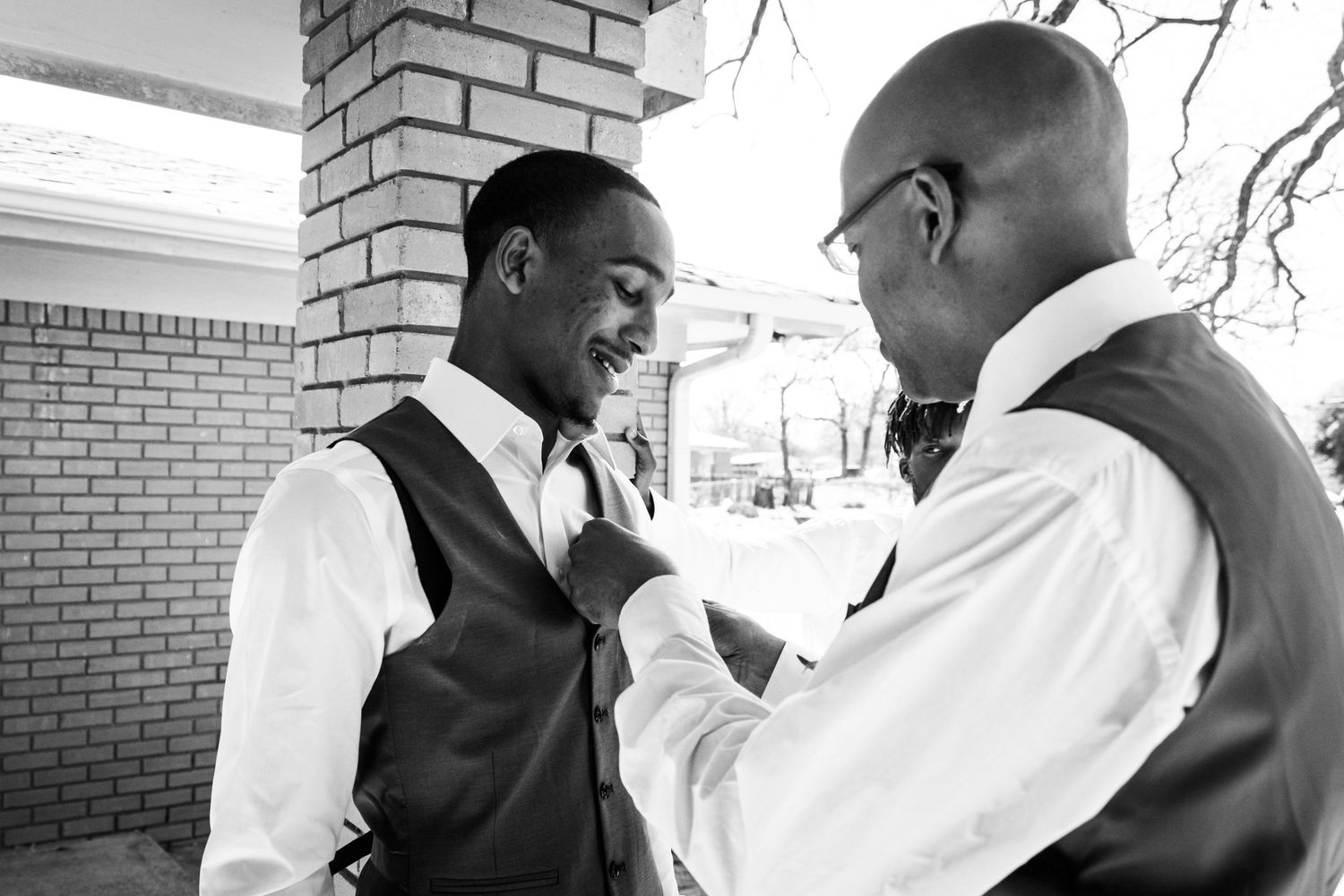Two men in formal attire helping each other with wedding preparations.