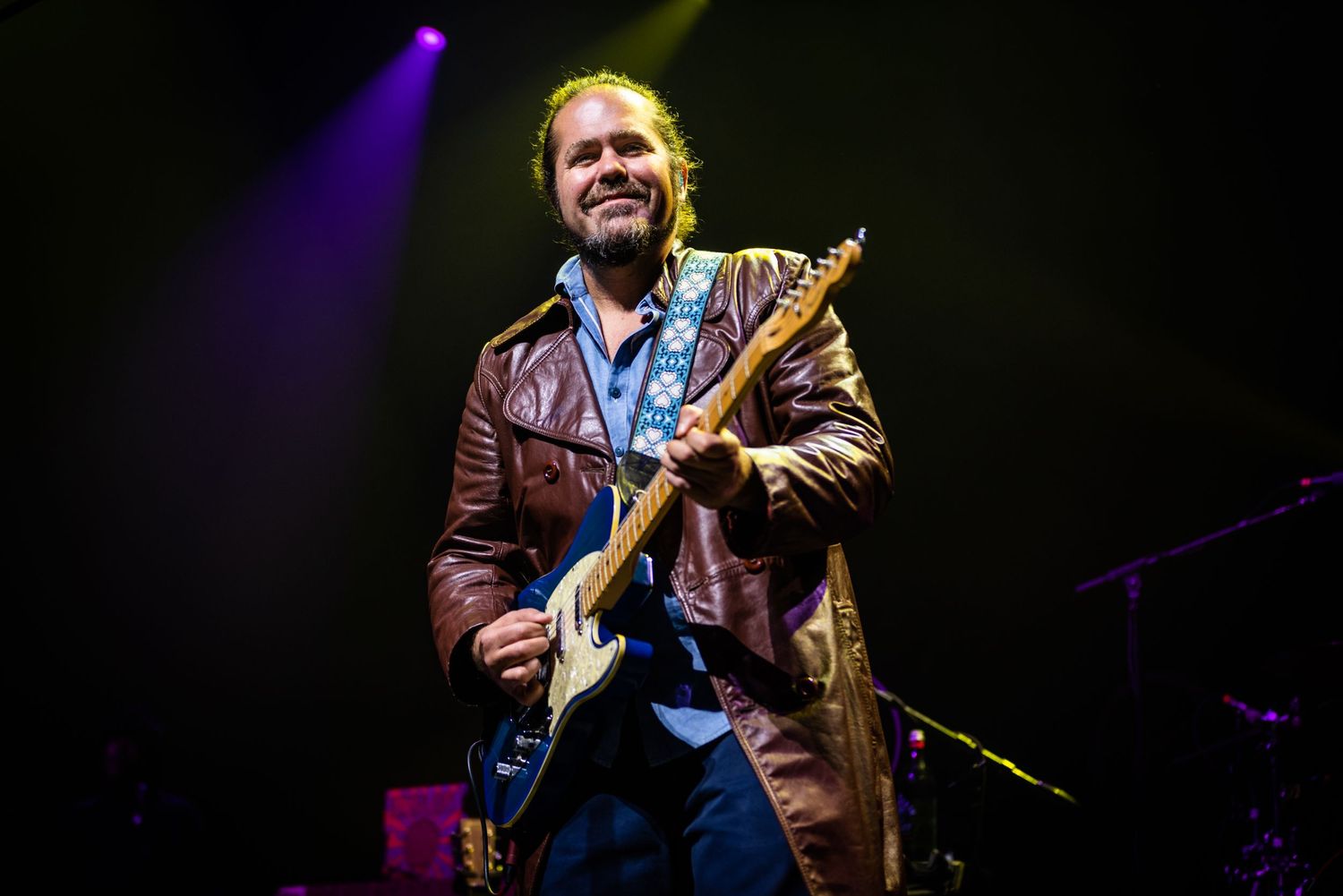 Musician in leather jacket plays electric guitar under dramatic stage lighting.