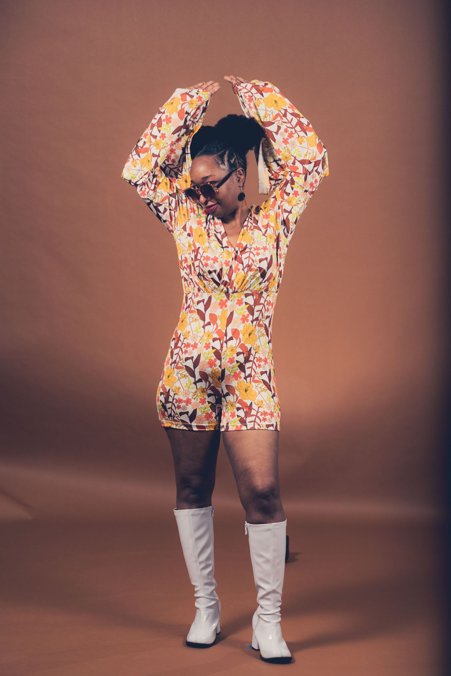 Person in floral romper and white boots striking dynamic pose against brown backdrop.