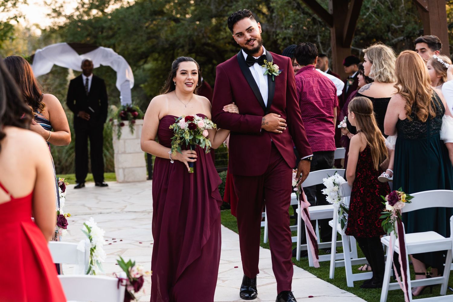 Wedding party members in burgundy attire walking down an outdoor ceremony aisle.