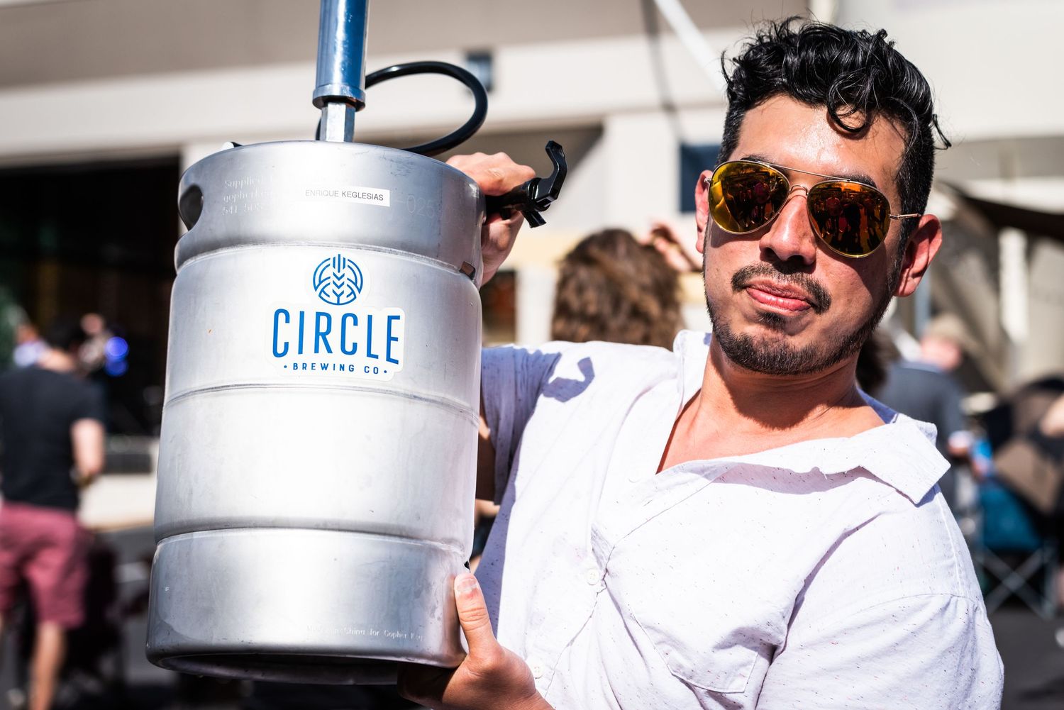 Person smiling while holding a Circle Brewing Company beer keg in bright sunlight.