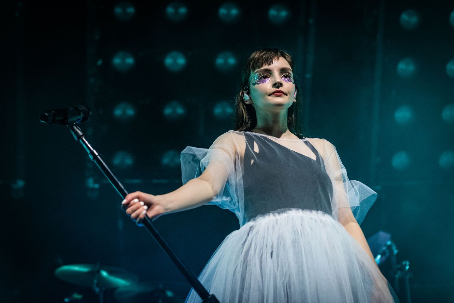Stage performer wearing a white tulle dress illuminated by blue stage lights during a concert.