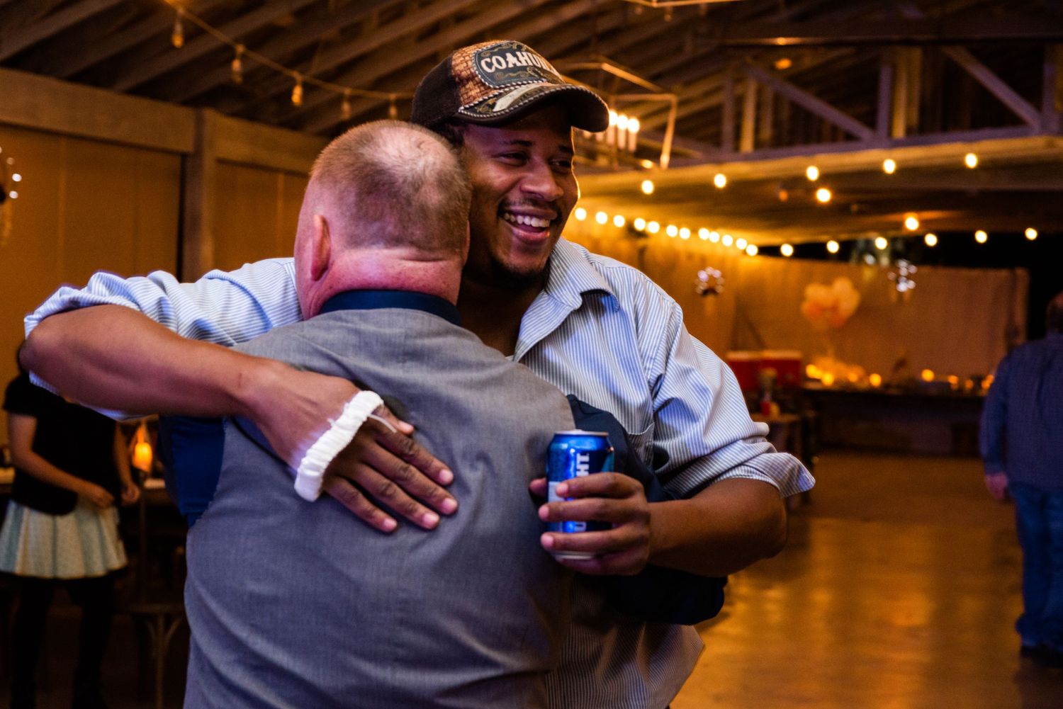 Two guests share a warm embrace while holding beverages at an indoor reception venue with string lights overhead.