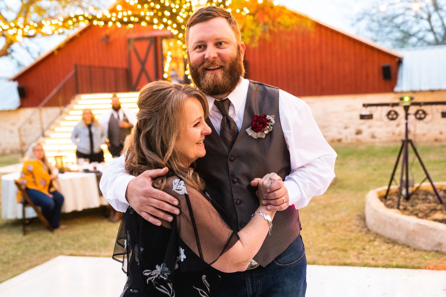 Couple shares intimate moment at rustic barn wedding reception with string lights decorating background.