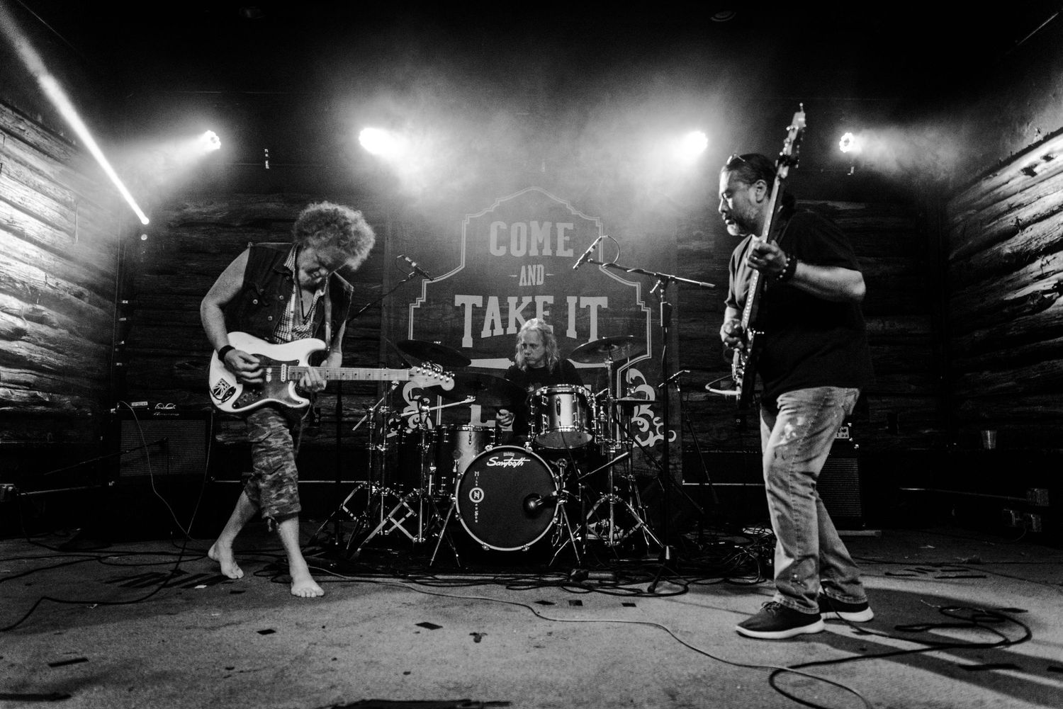 Wide shot of band performing on dark stage with drums and guitar players visible.