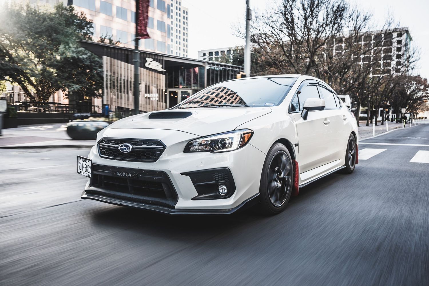 Modified white Subaru WRX STI with red brake calipers captured in dynamic motion on urban road.