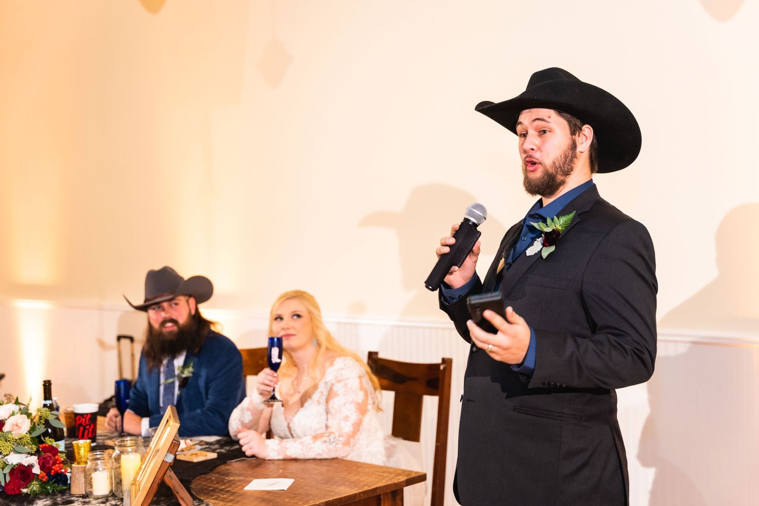 A speaker in a black cowboy hat and suit addresses wedding guests during a reception.