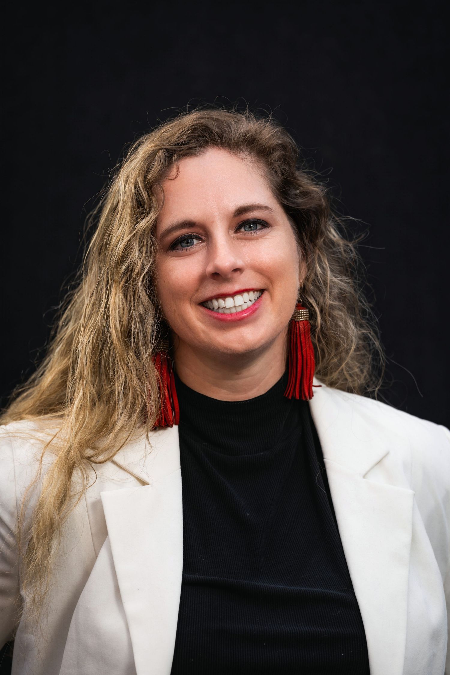 Stylish portrait of woman in white blazer with wavy hair and red tassel earrings.