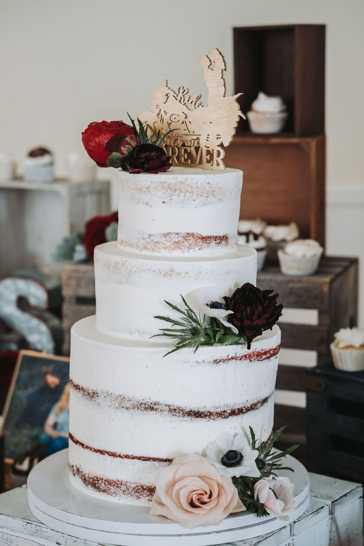Three-tiered semi-naked wedding cake decorated with burgundy flowers and wooden topper.