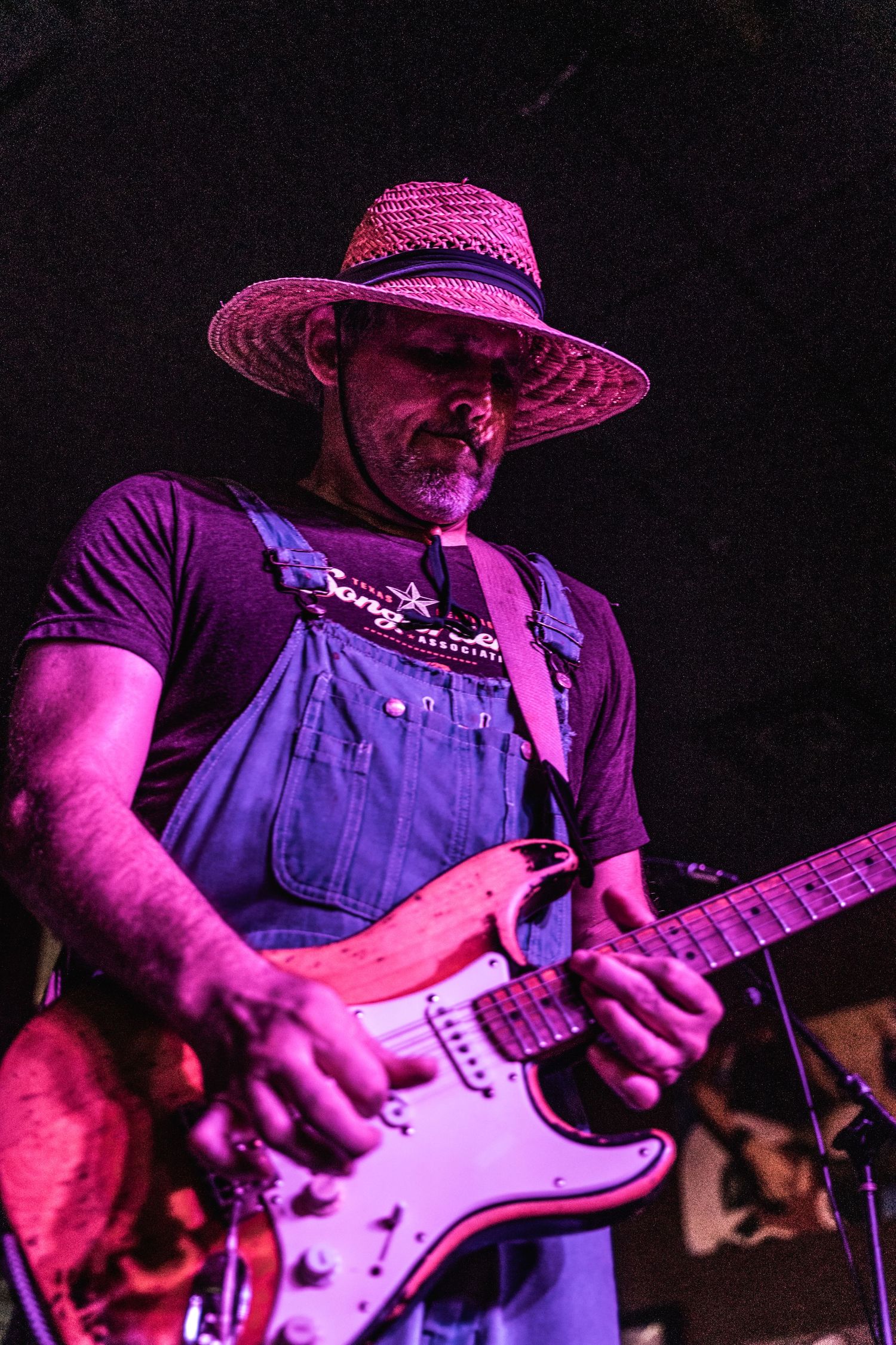 Performer in straw hat and overalls plays Stratocaster guitar under magenta lighting.