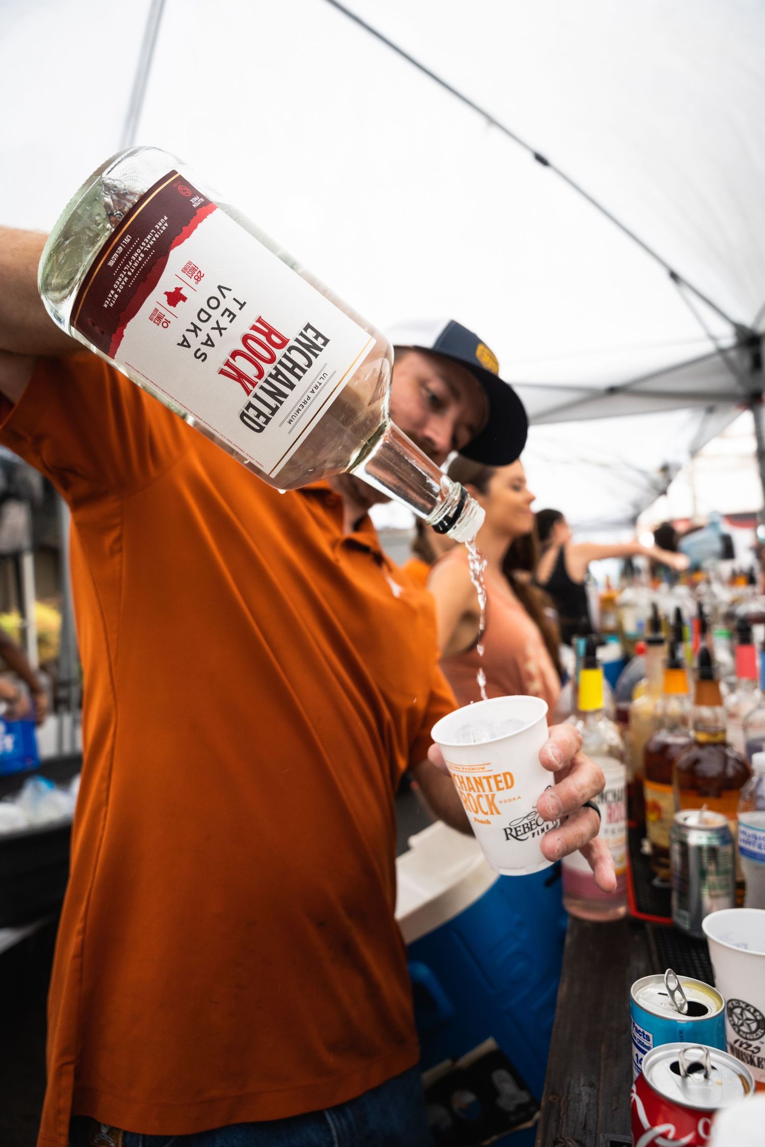 Person in orange shirt pouring spirits from bottle into cup under white tent.