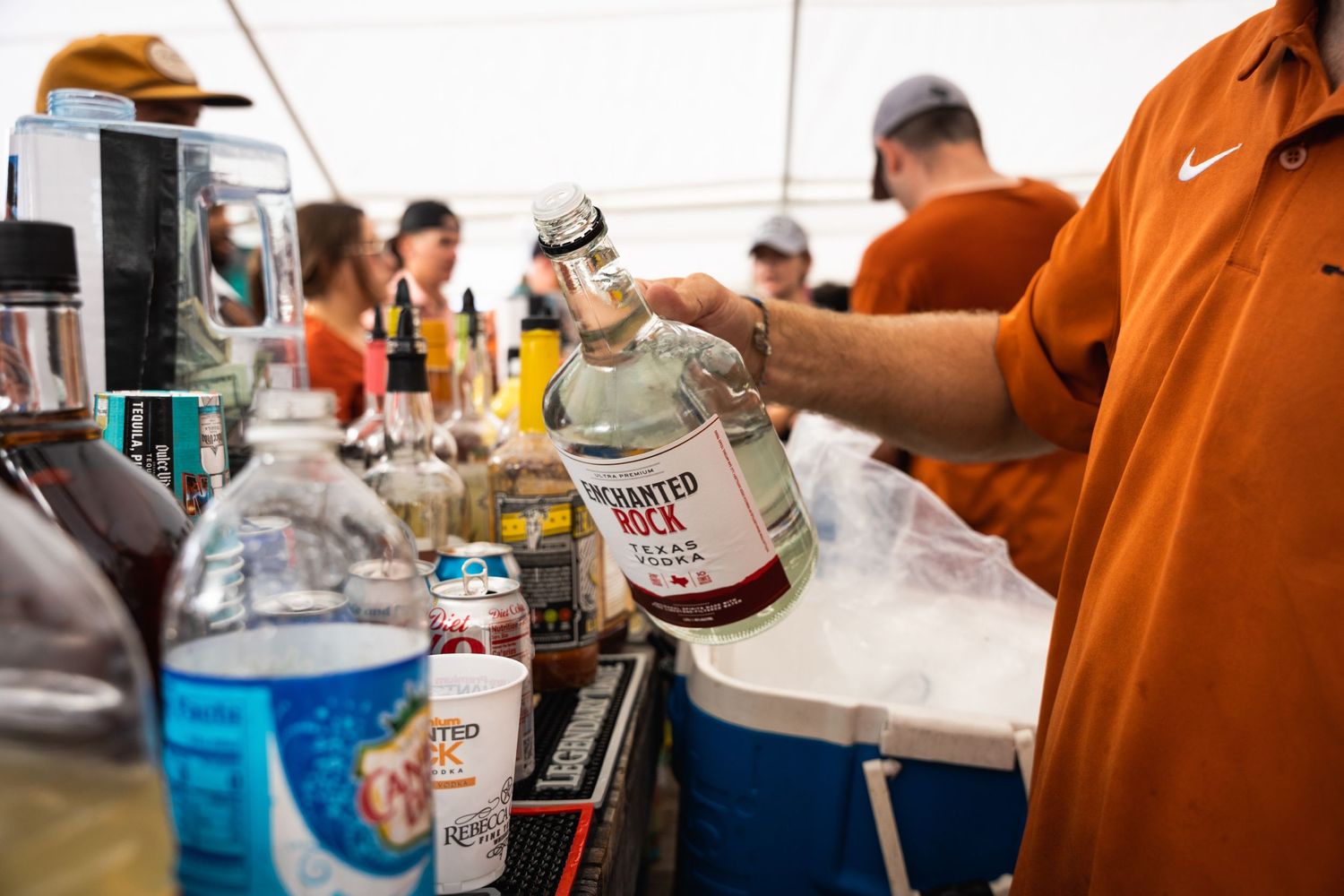 Hands pour drinks from bottles at an outdoor bar setup with coolers.