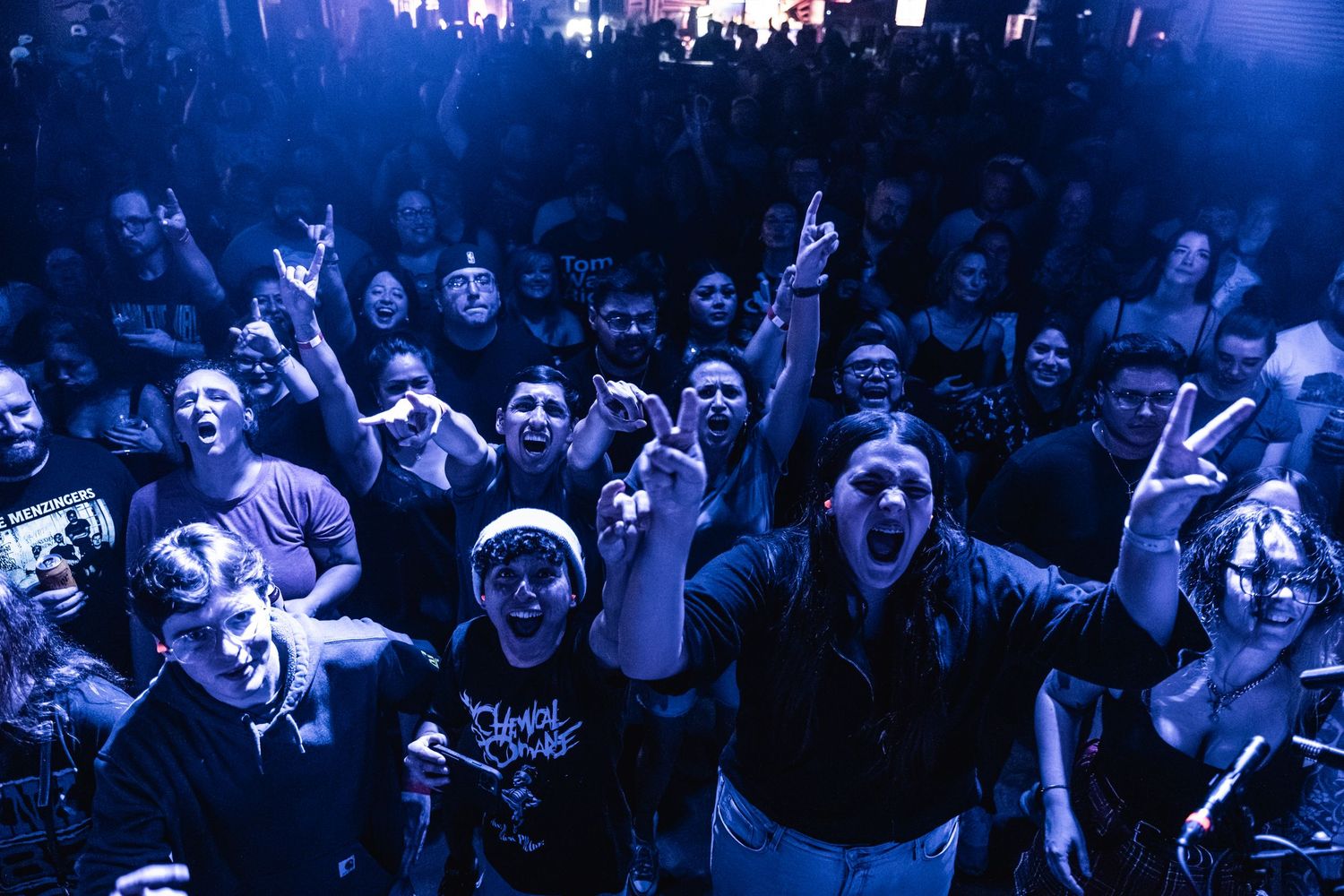 Enthusiastic crowd of concertgoers cheers and raises hands at a music event bathed in blue stage lighting.