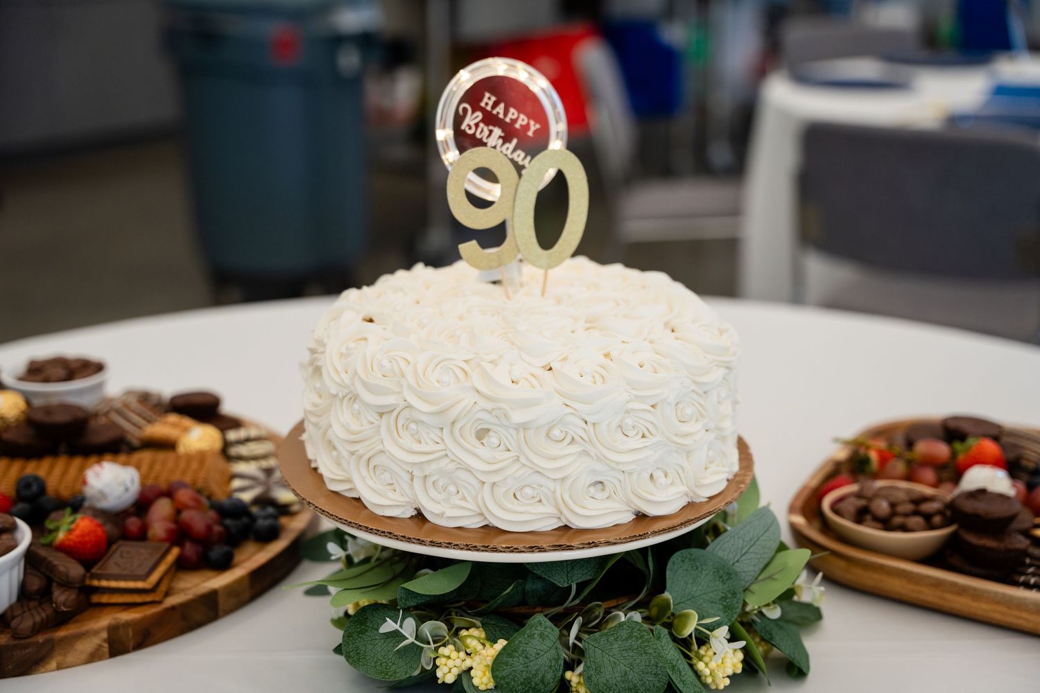 White frosted celebration cake with gold number 90 topper on green leaf garland.