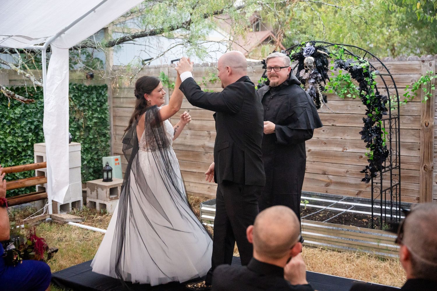 A wedding ceremony takes place under a white canopy with an arch decorated with greenery.