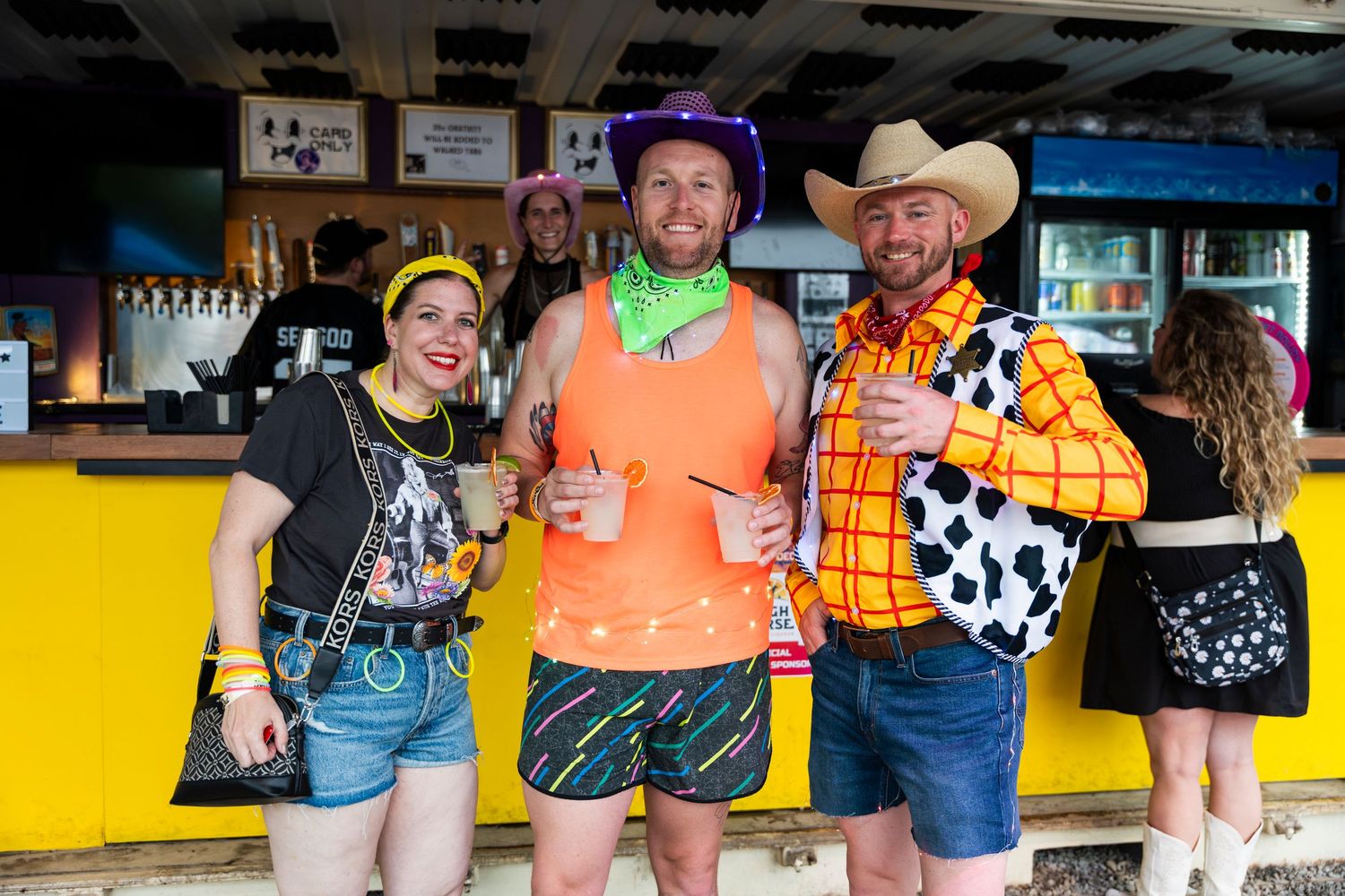 Group dressed in casual attire with western and cowboy-themed outfits pose together at a bar counter.