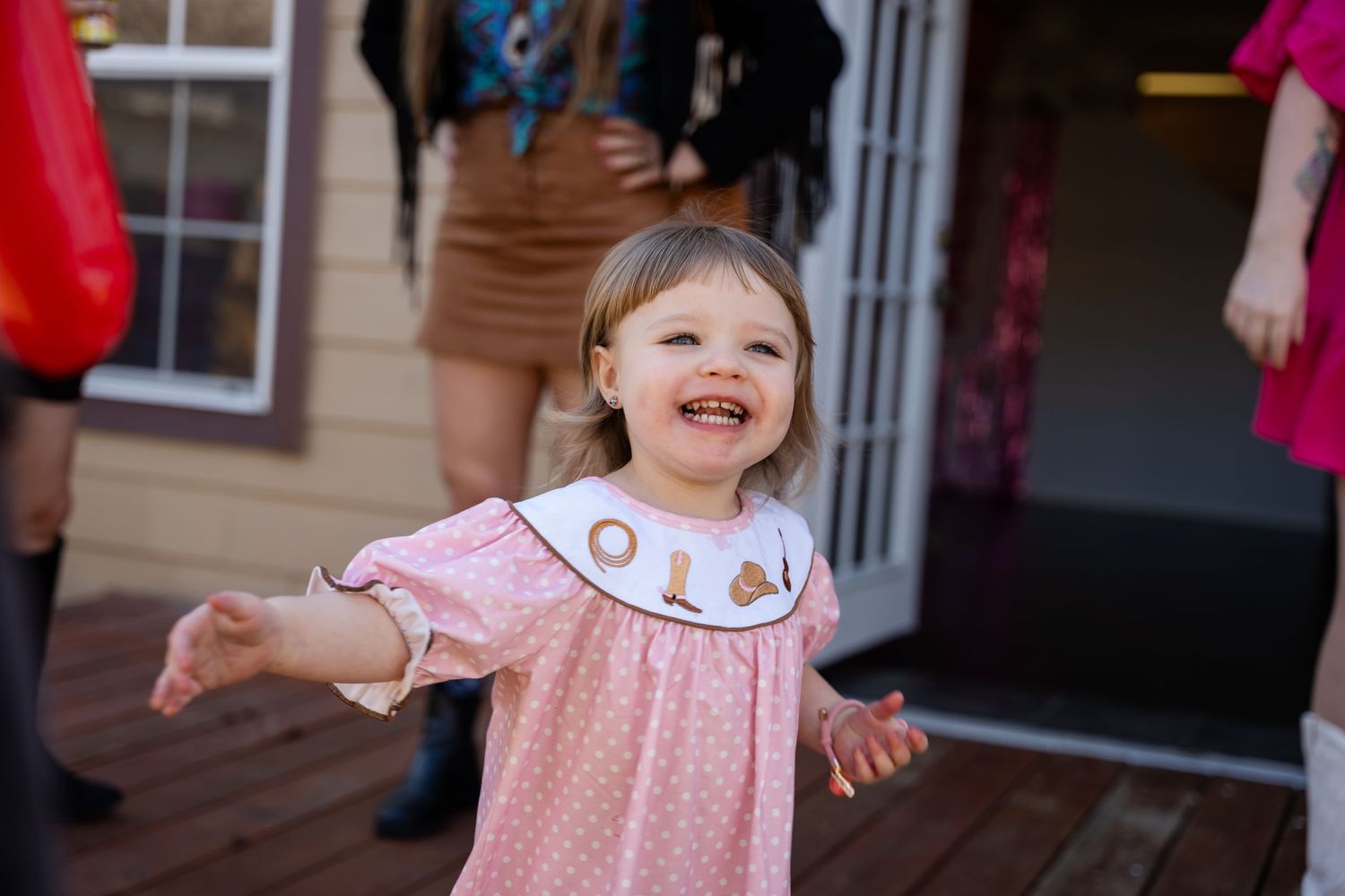 A toddler in a pink polka dot dress spreads their arms joyfully while standing on a wooden deck.