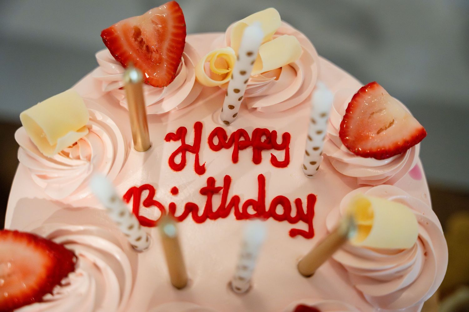 A pink birthday cake decorated with whipped cream swirls, fresh strawberries and white chocolate pieces featuring red lettering.