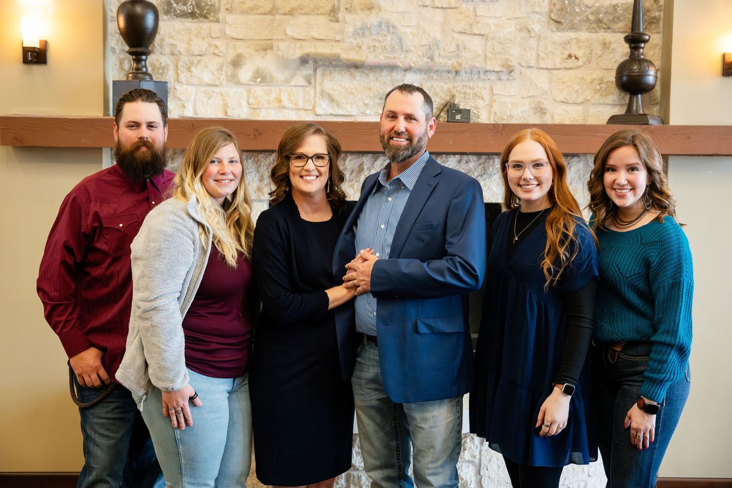 A group of six people in business casual attire stand together smiling against a stone wall with warm lighting.