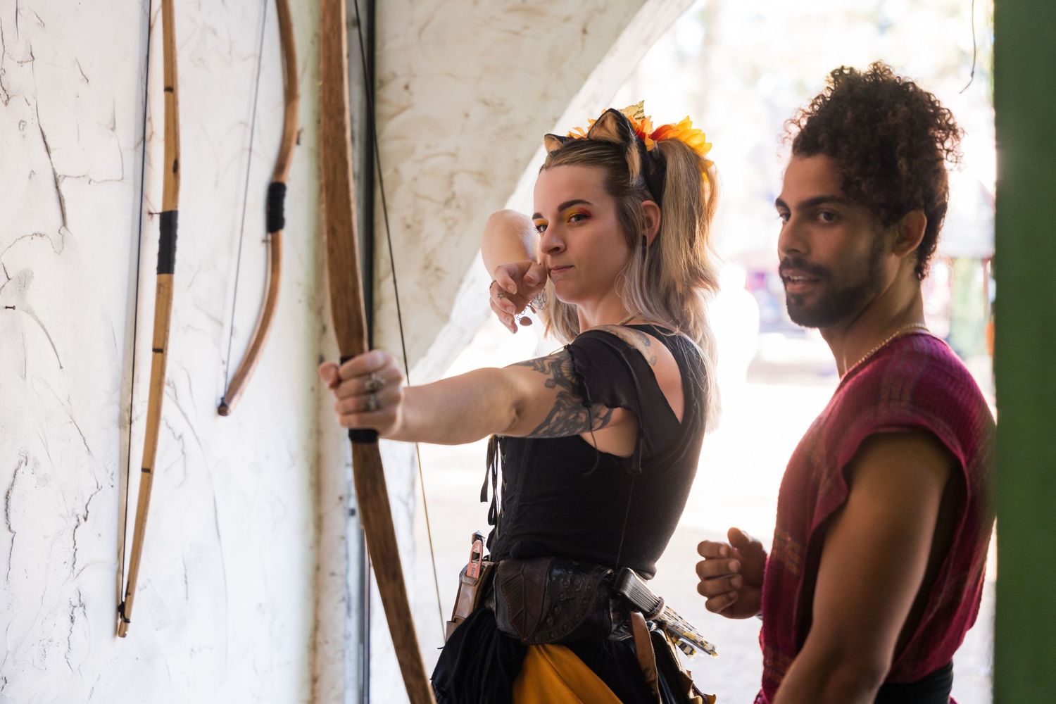 Two performers examine archery equipment against a white wall.