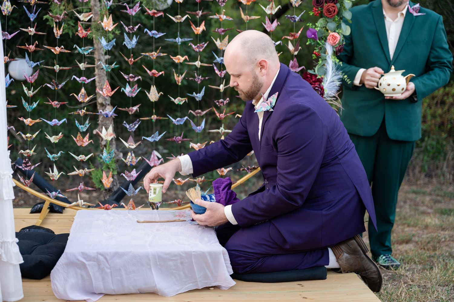 Person in purple suit kneels at ceremonial table with origami crane backdrop.