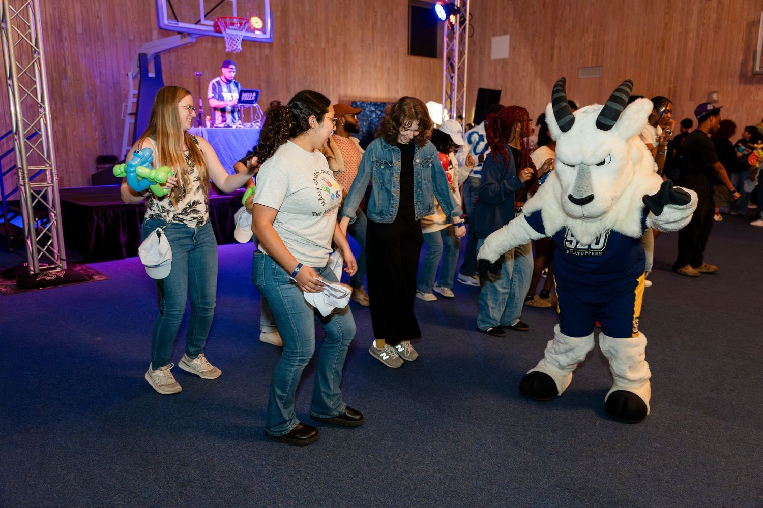 People dancing at an indoor event with a sports mascot in white and blue joining the fun under colored lighting.