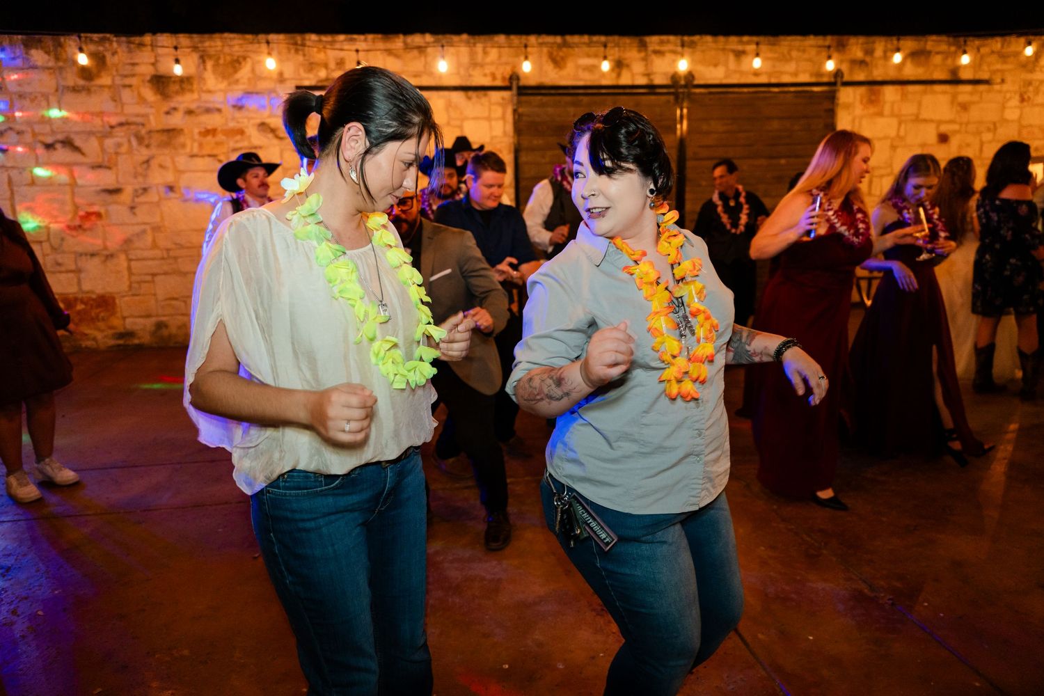 Party guests wearing Hawaiian leis dance under string lights at an indoor venue.