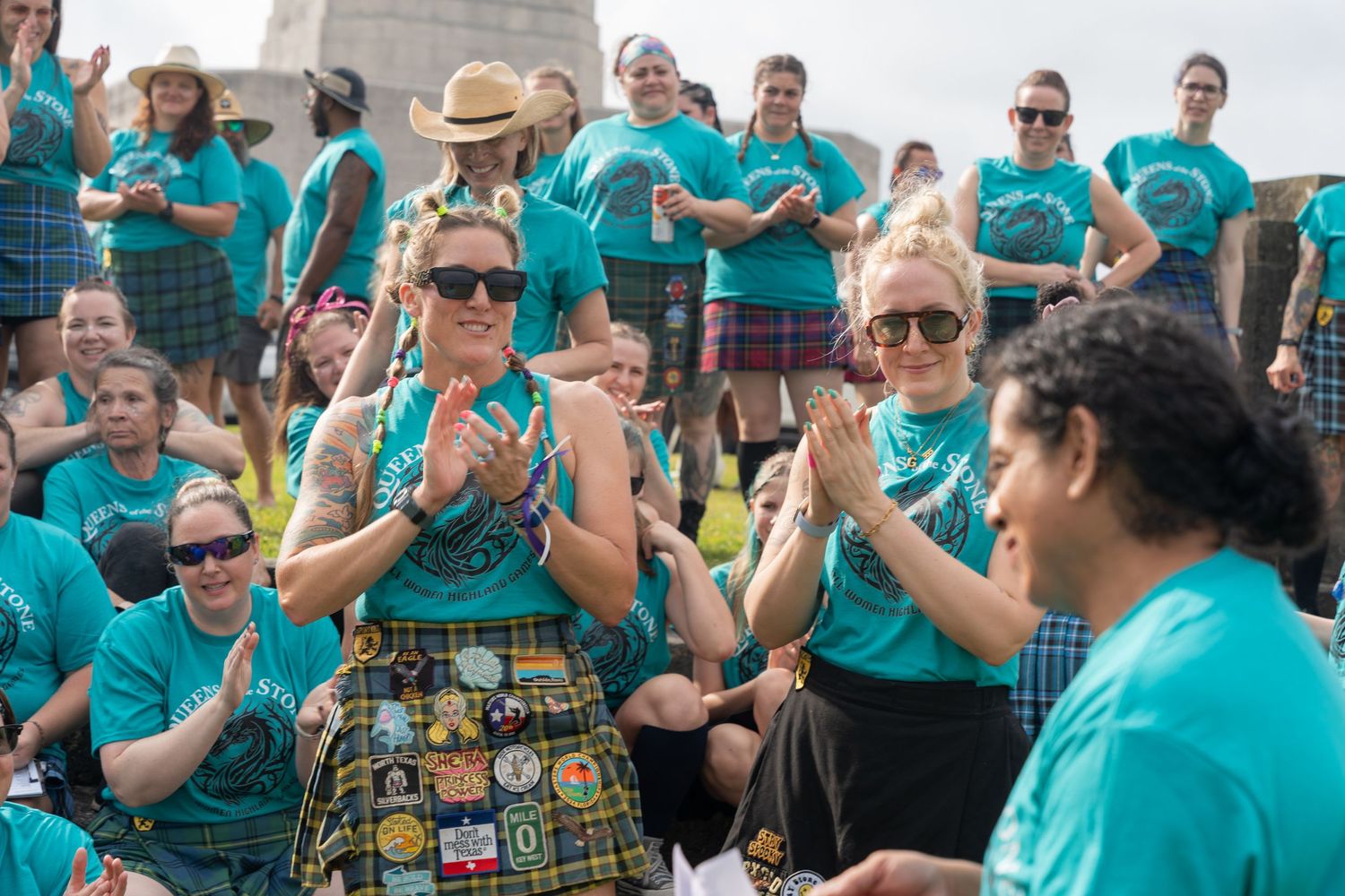 Group of women in teal shirts and kilts clapping at an outdoor event near a monument.
