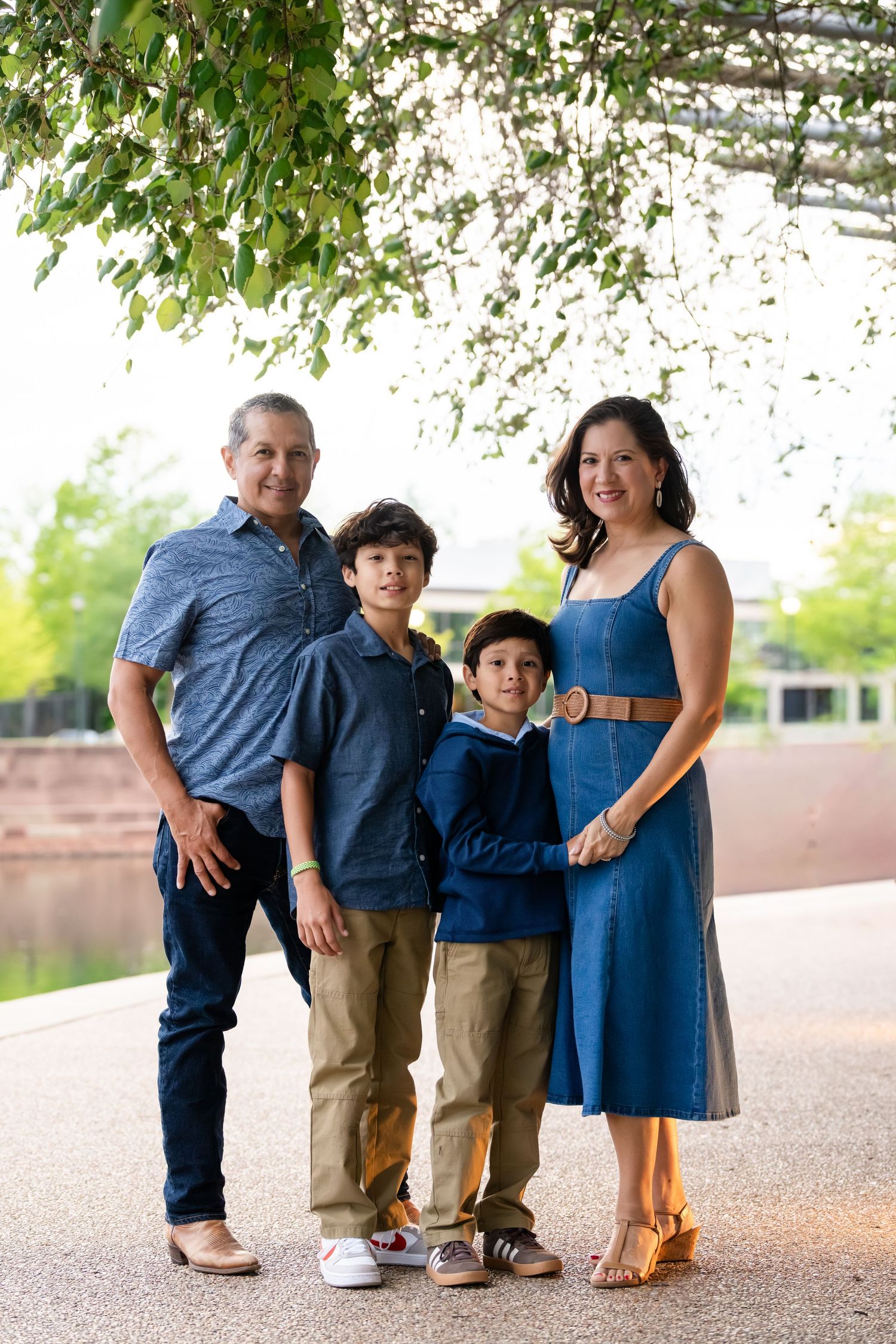 Family of four posing outdoors under hanging vines near a pond, wearing coordinated blue and tan outfits.