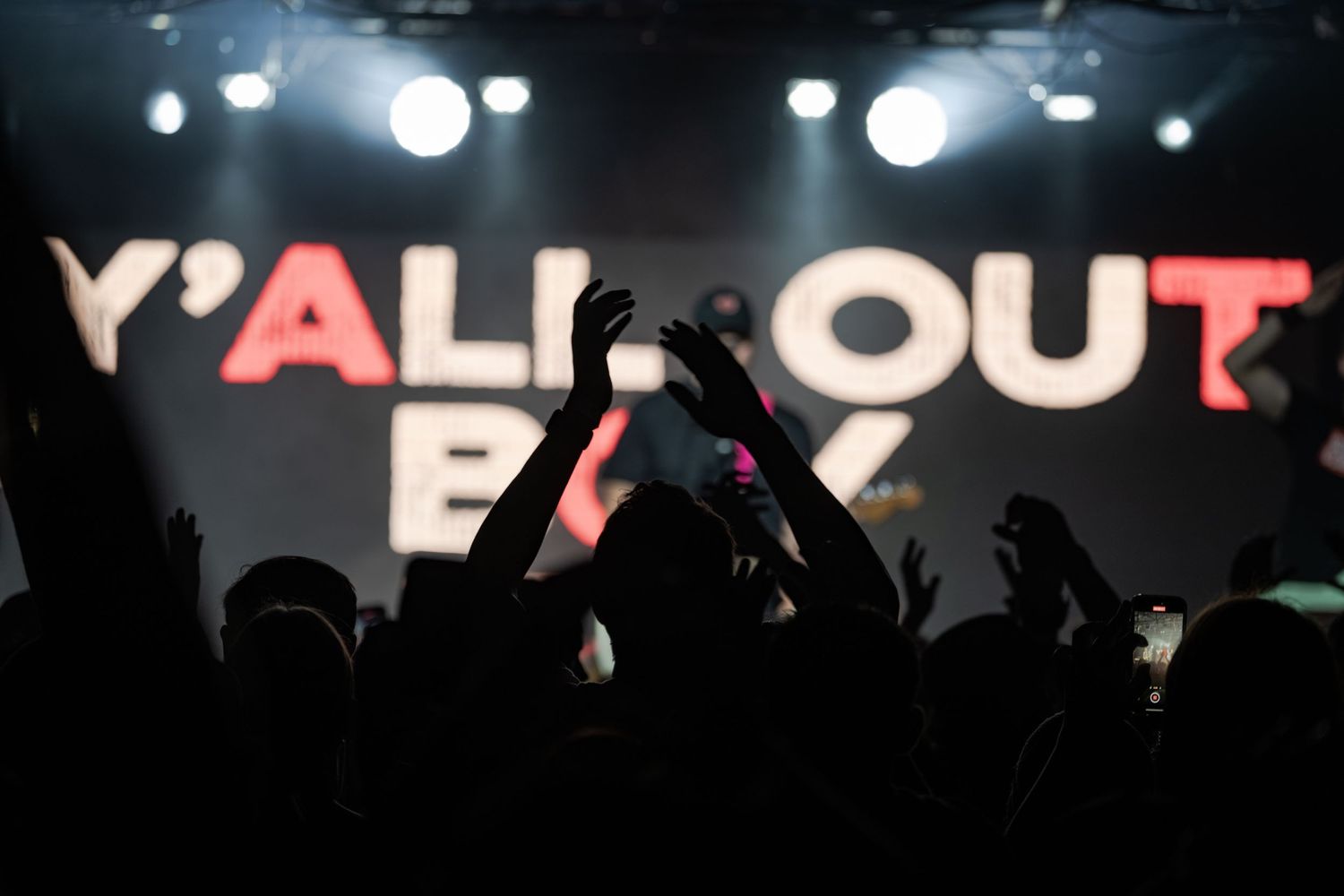 Crowd silhouettes raise hands at a Y'All Out Boy concert with bright stage lighting illuminating the venue.