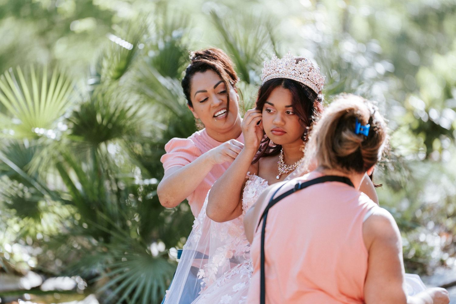 Woman in tiara gets necklace adjusted by helper outdoors near tropical palms during a formal event.