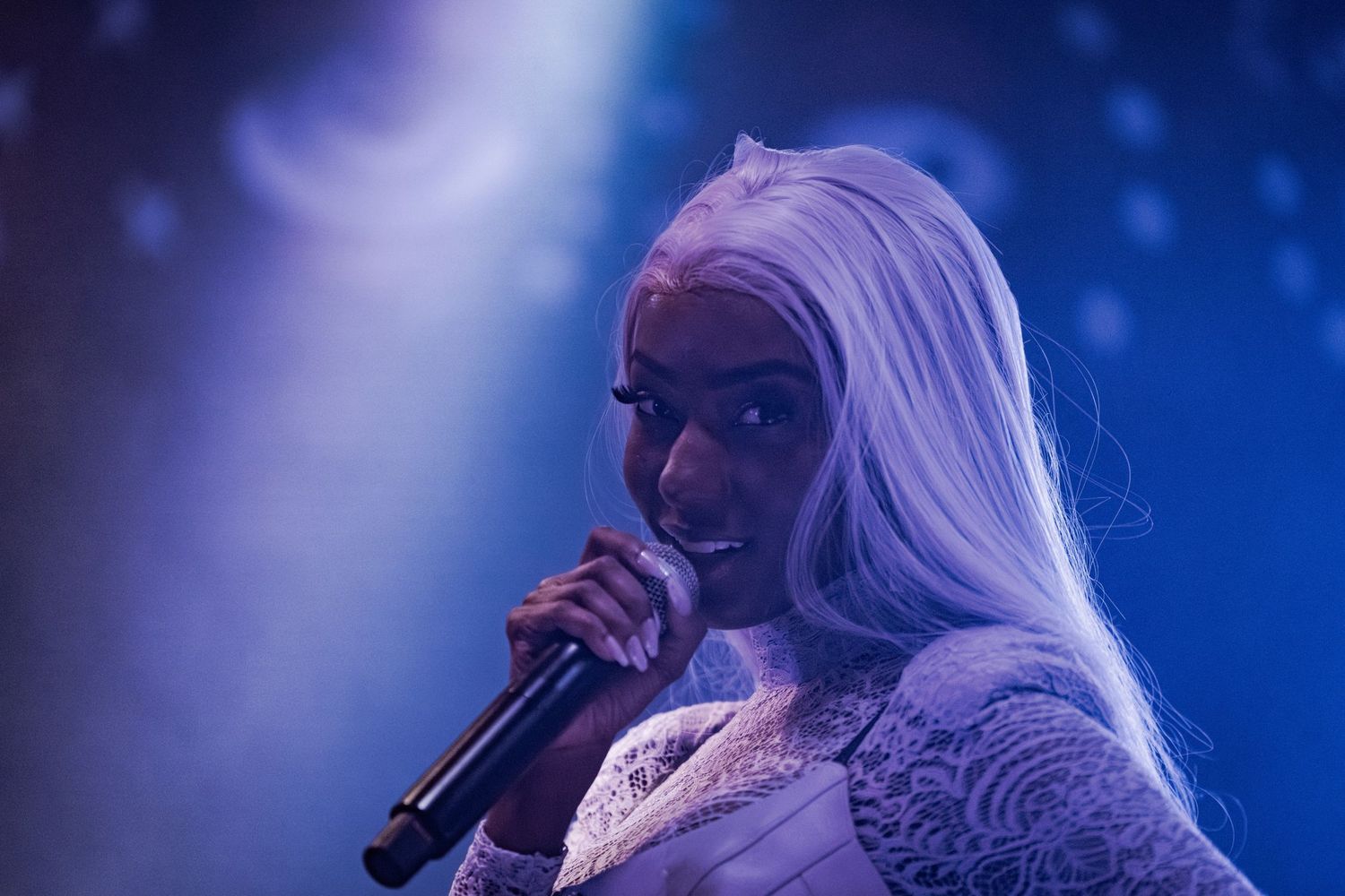 Black female singer with white hair performing on stage, holding microphone under blue spotlight in white lace outfit.
